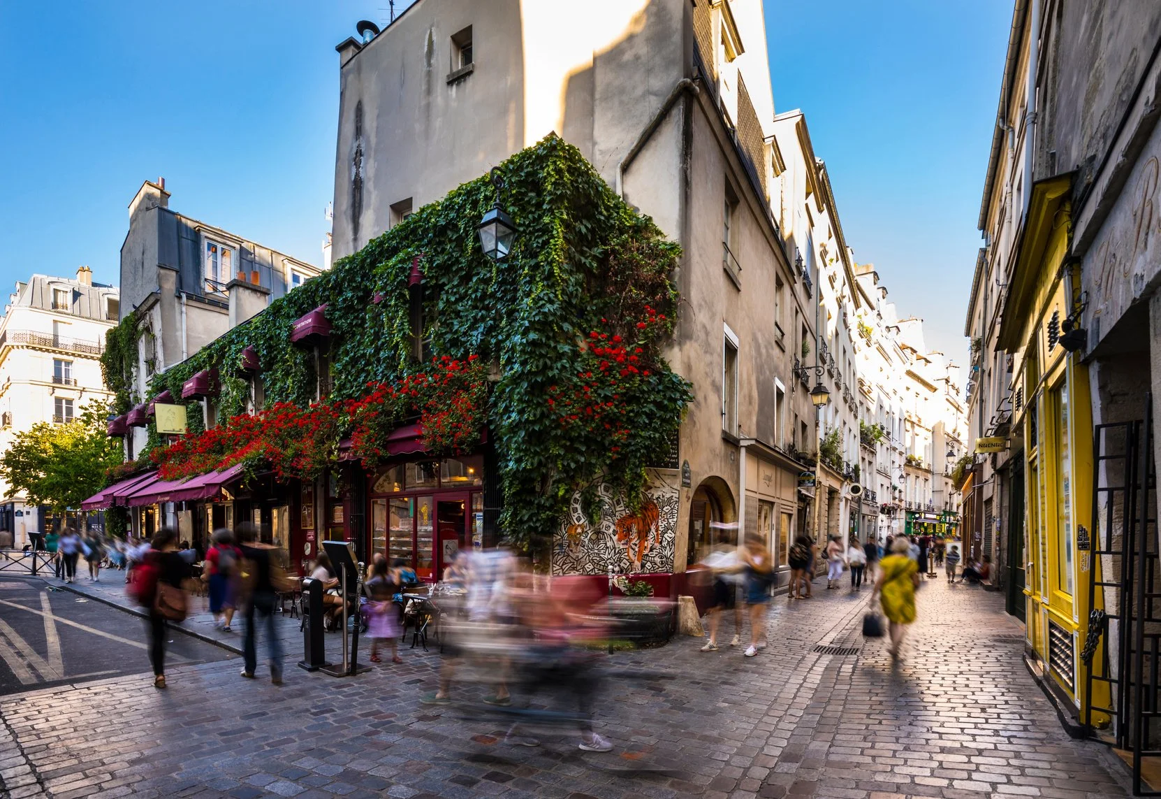 A busy Paris street scene with people walking on cobblestone pavement in front of a corner building covered in green ivy and red flowers, with cafes and shops along the street.