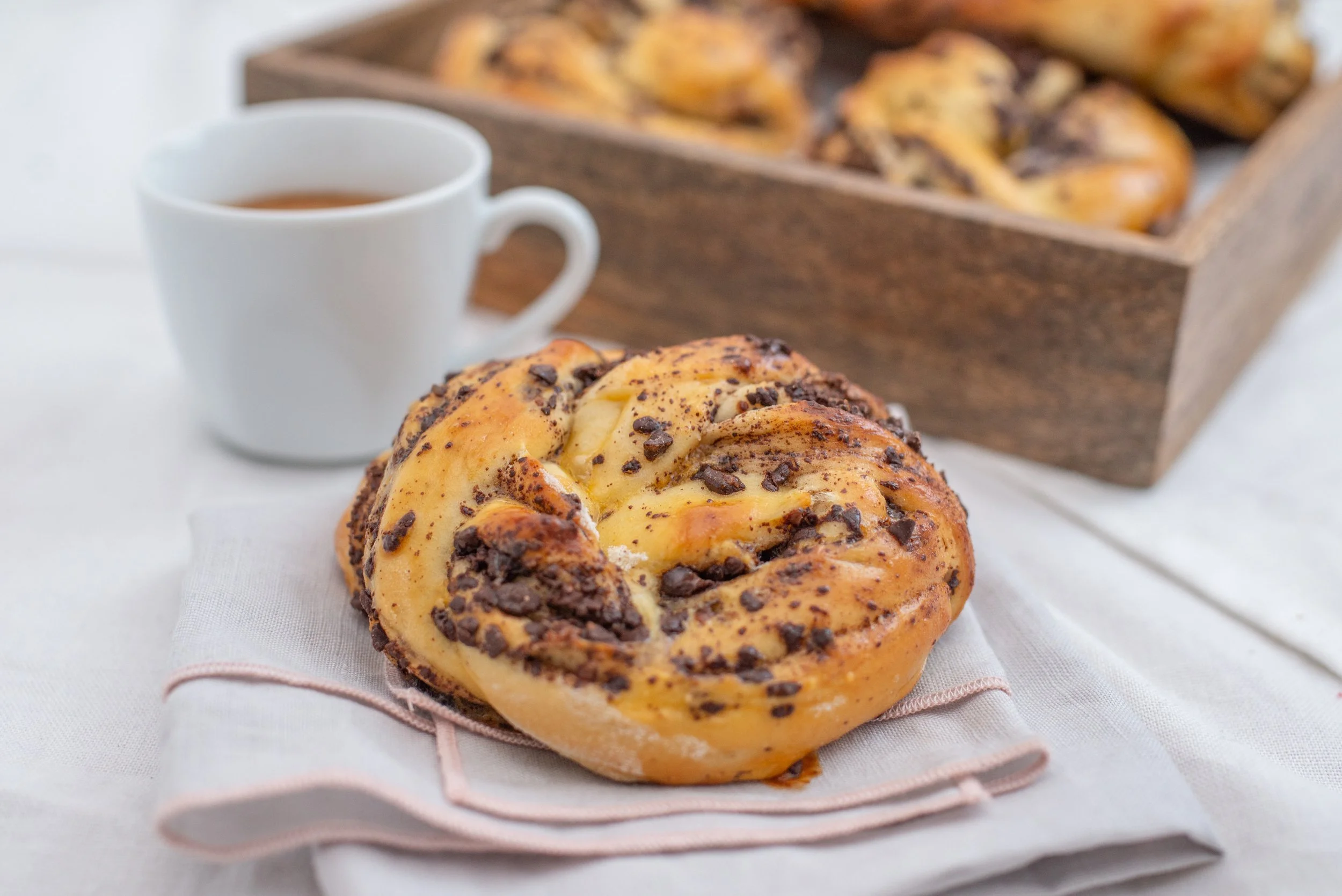 A close-up of a chocolate chip cinnamon roll on a napkin, with a white cup of coffee or tea and a wooden tray of more cinnamon rolls in the background.