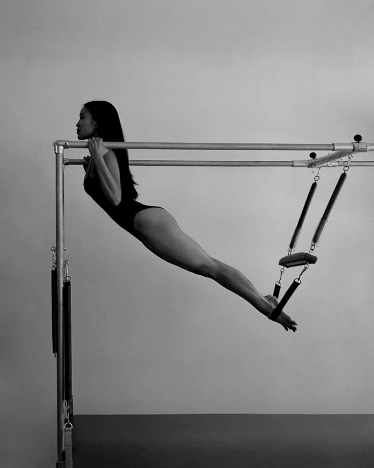 A woman is performing a Pilates exercise on a trapeze table, holding a pull-up position, using the apparatus for support.
