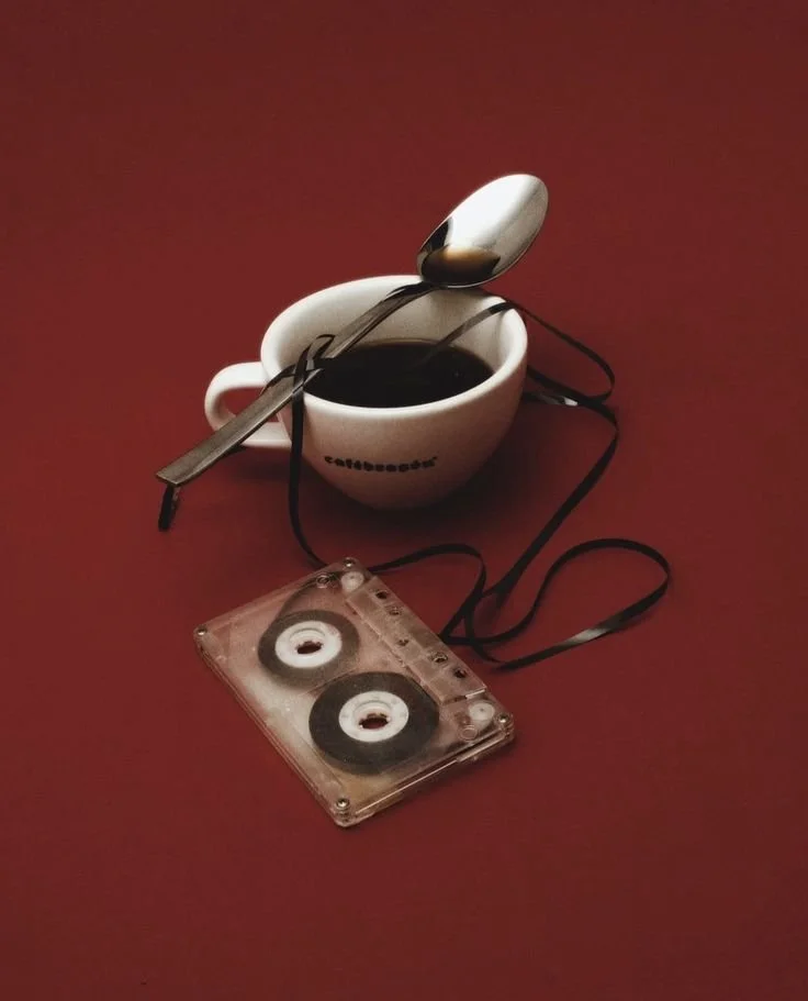 A white coffee cup filled with dark coffee, a metal spoon resting across the cup, and a transparent tape recorder with tape reels, all on a red surface.