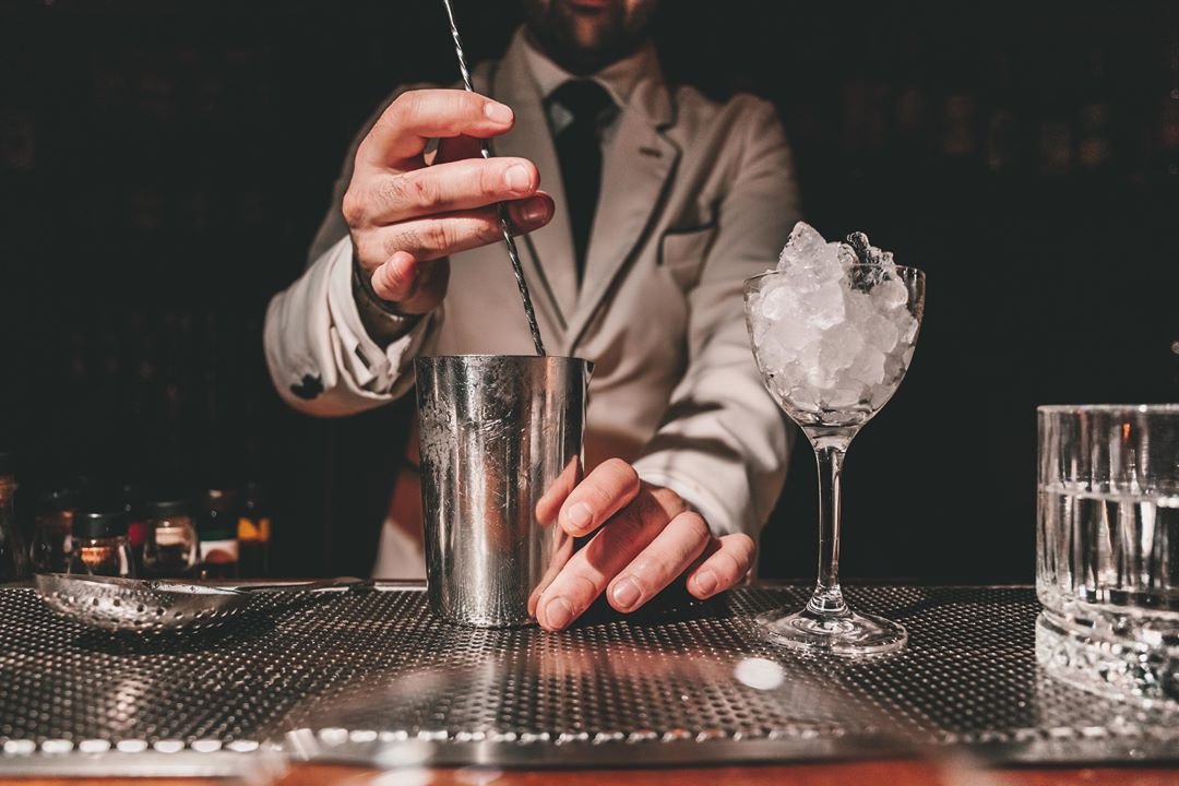 A bartender in a beige suit preparing a cocktail with a shaker, spoon, and ice in a martini glass behind the bar.