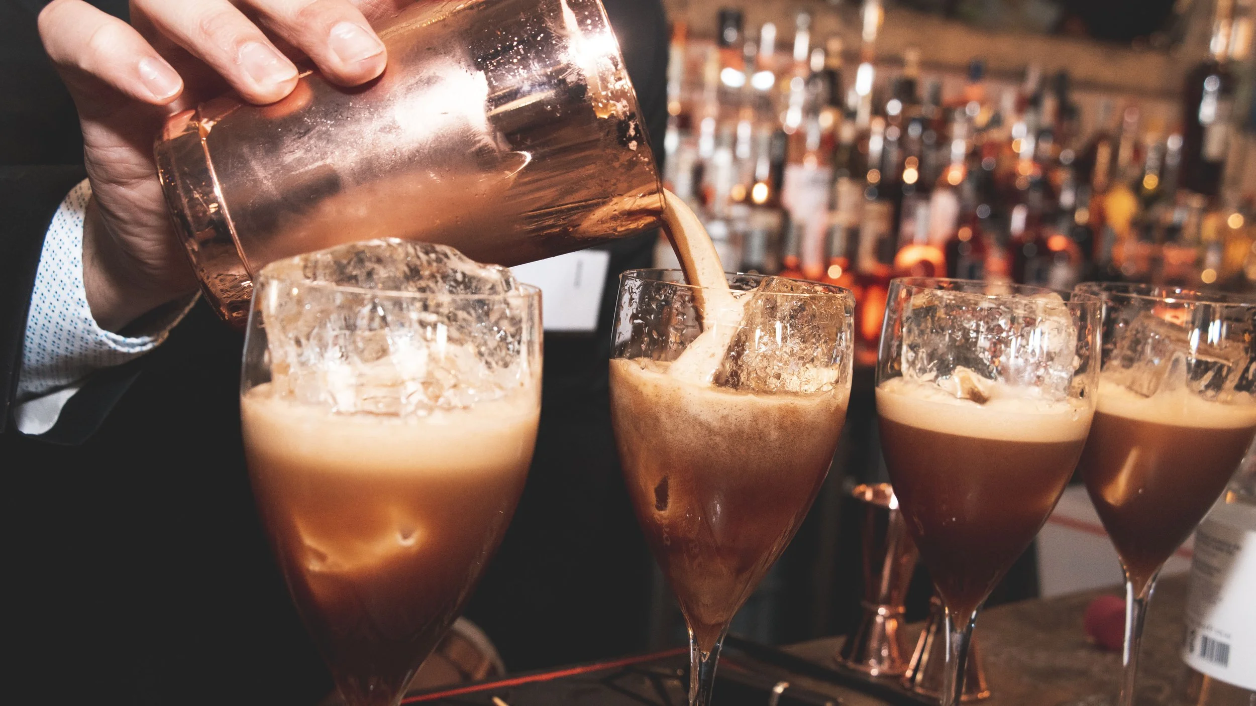 A bartender pours a creamy brown cocktail from a copper shaker into tall glasses filled with ice, with a bar filled with various bottles in the background.