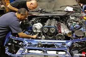 Two men working on a car engine, with engine parts and tools visible under the hood.