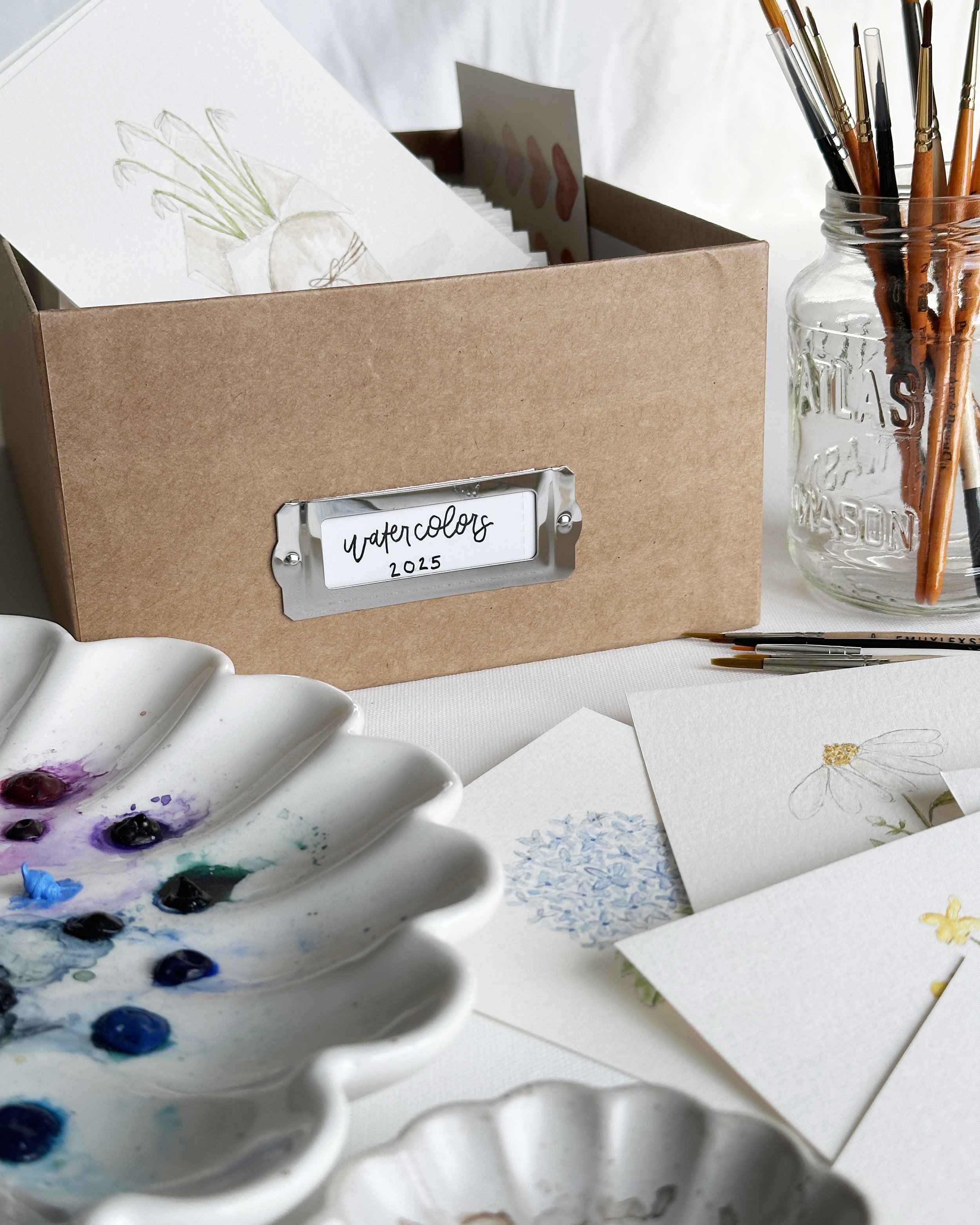 A photo of watercolor brushes, paint palette, and small paintings on a studio table.