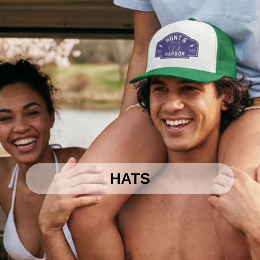 Two smiling friends, a woman and a man, enjoying time outdoors near a body of water. The man is wearing a green and white hat with a patch reading "Hunt & Harbor." The woman's hair is curly and she is wearing a white top. The man is shirtless, and th
