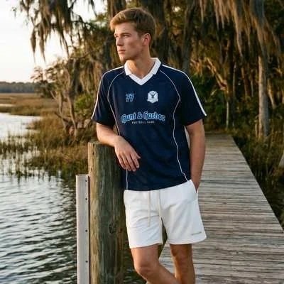 A young man in a navy soccer jersey and white shorts standing on a wooden dock near water, with trees in the background.