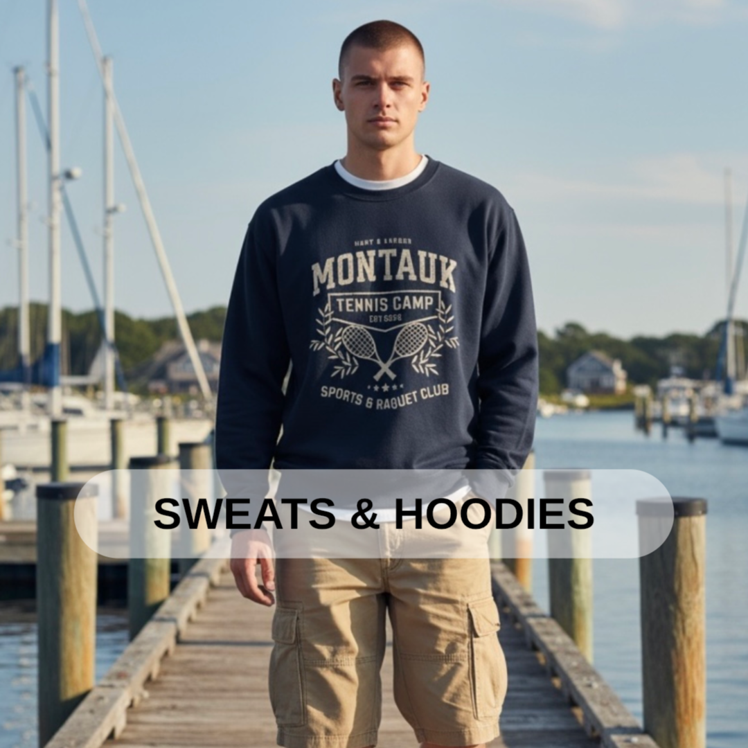 A young man standing on a wooden dock near boats with water and houses in the background, wearing a navy sweatshirt with white text and beige cargo shorts, with the overlay text 'SWEATS & HOODIES'.