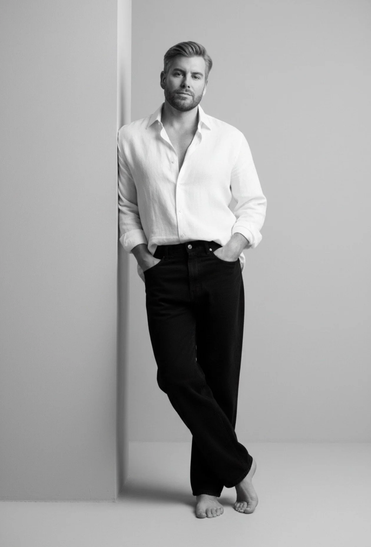 Black and white photo of a man standing barefoot against a plain wall, wearing a white dress shirt and dark jeans, with a relaxed pose and confident expression.
