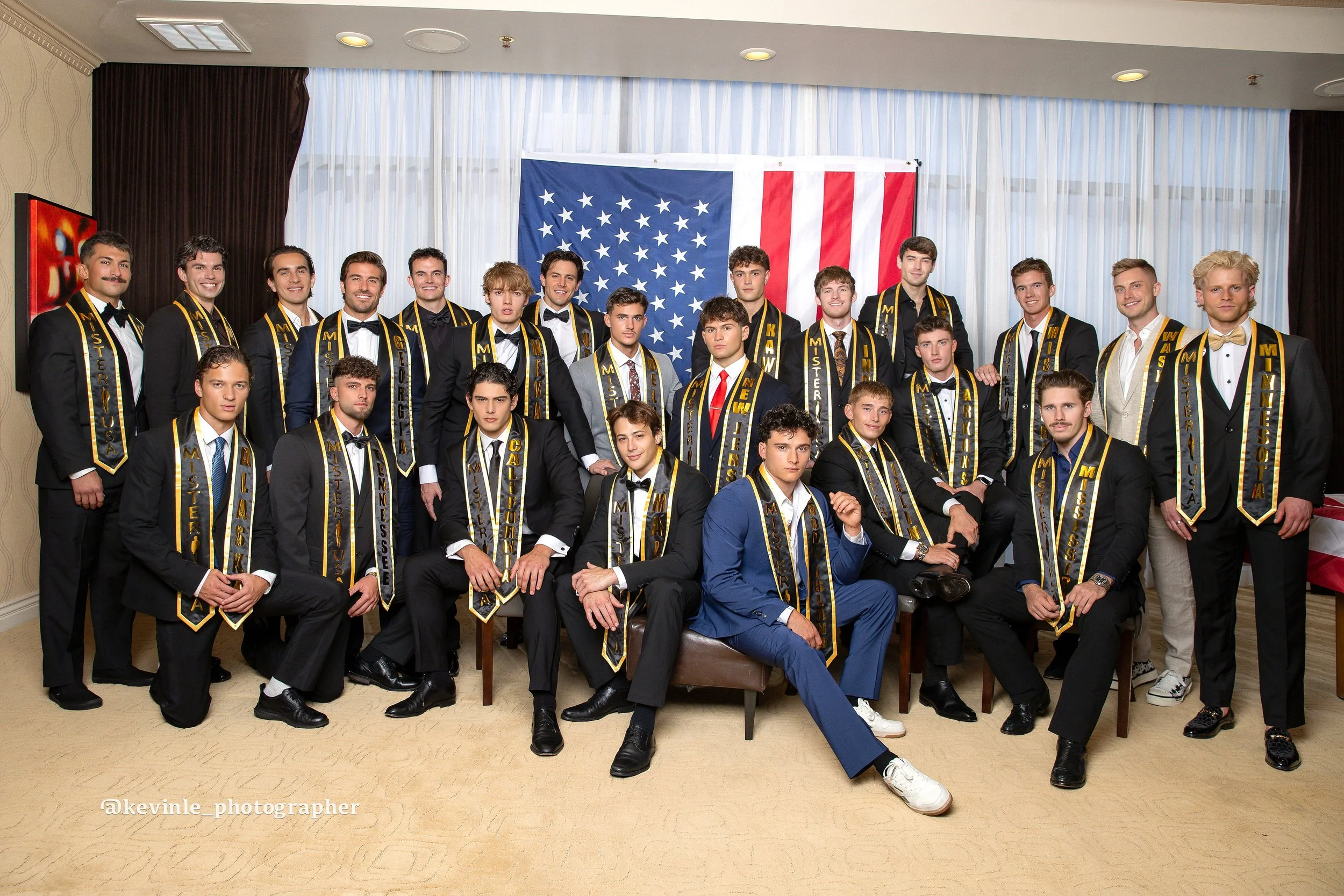 Mister USA 2026 contestants in Las Vegas. A large group of young men dressed in formal tuxedos and sashes, posing for a photo in front of an American flag, during Mister USA finals in Las Vegas.