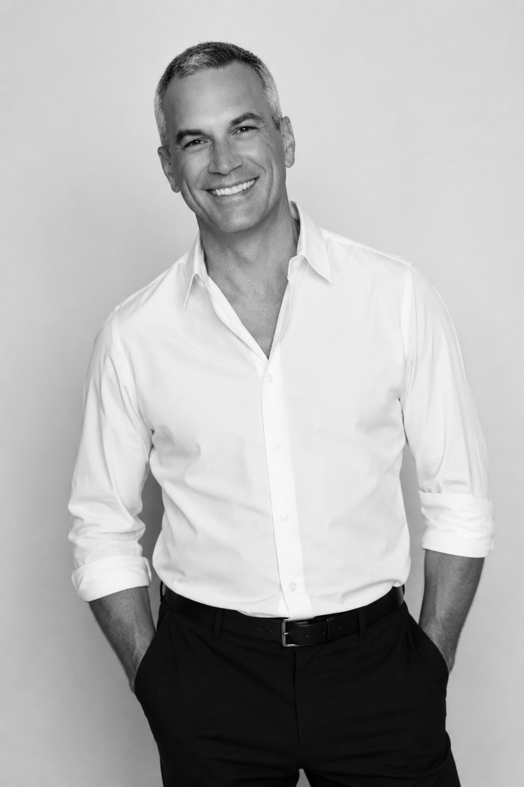 Black and white portrait of a smiling man with short gray hair wearing a white button-up shirt with sleeves rolled up and black pants.