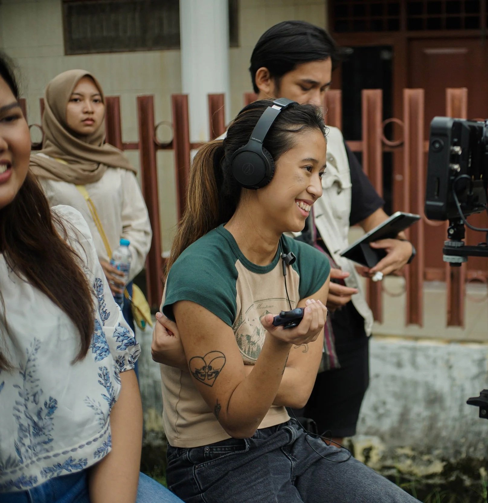 A woman wearing large black headphones, holding a remote control, smiling while sitting outdoors with a group of people. Others are standing nearby, some holding water bottles or electronic devices, and there is a camera on a tripod in front of her.