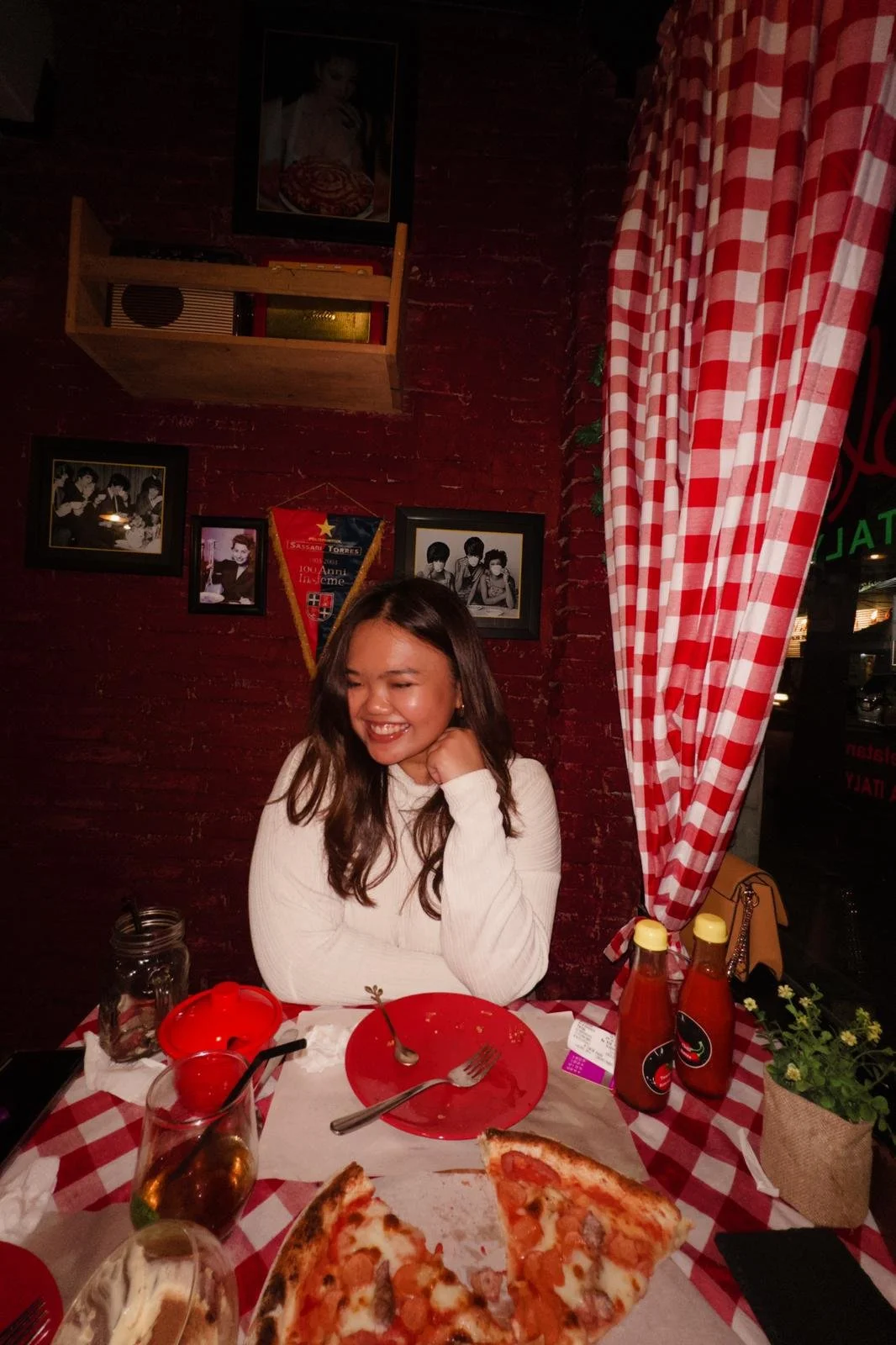 A woman is sitting at a table in a restaurant with a smile, wearing a white sweater. There are two slices of pizza, a red plate, a fork, a spoon, ketchup bottles, and a drink on the table. The background includes a brick wall with framed pictures, a red and white checkered curtain, a wooden shelf, and a floral arrangement.