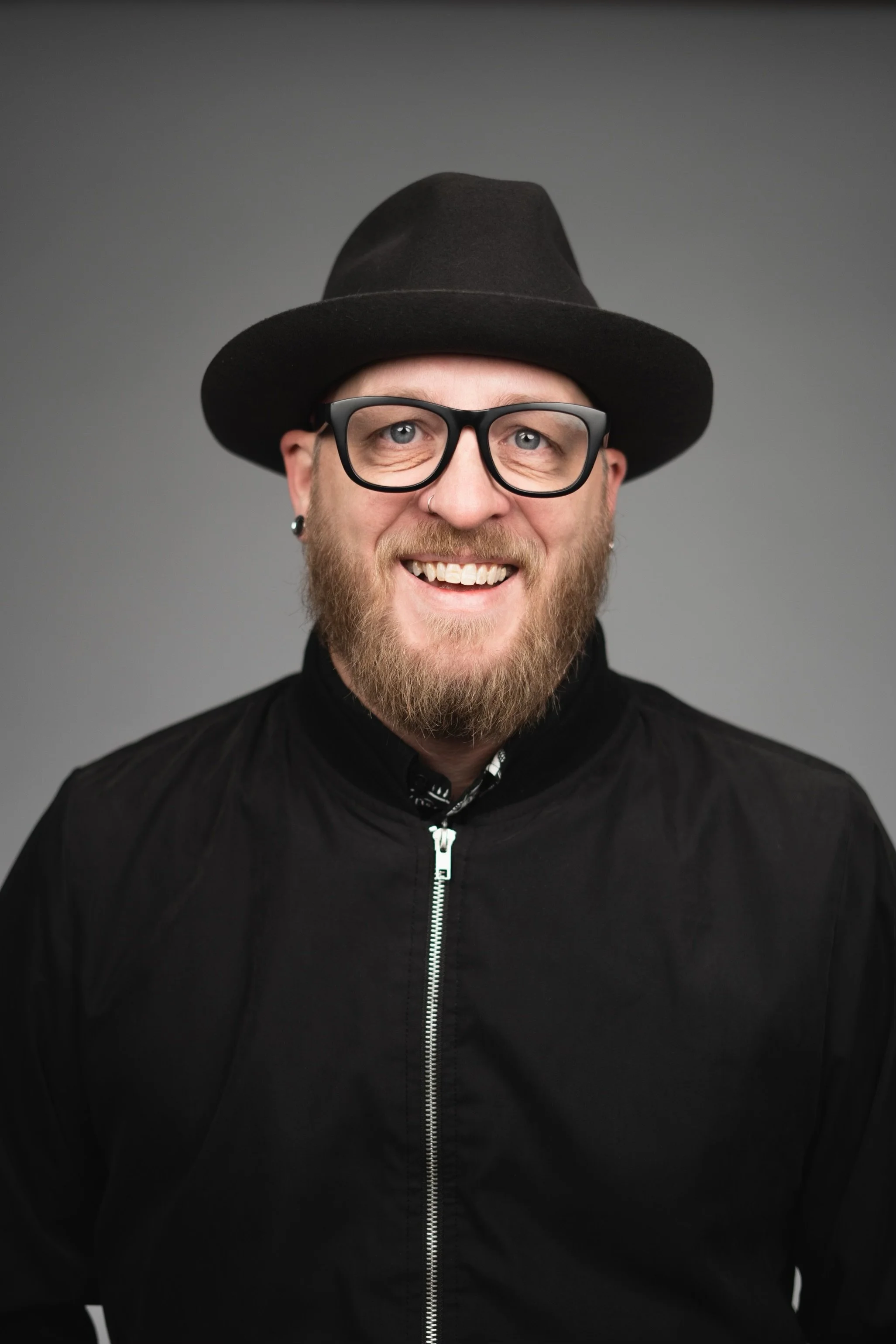A man with glasses and a beard wearing a black hat and black jacket, smiling against a gray background.