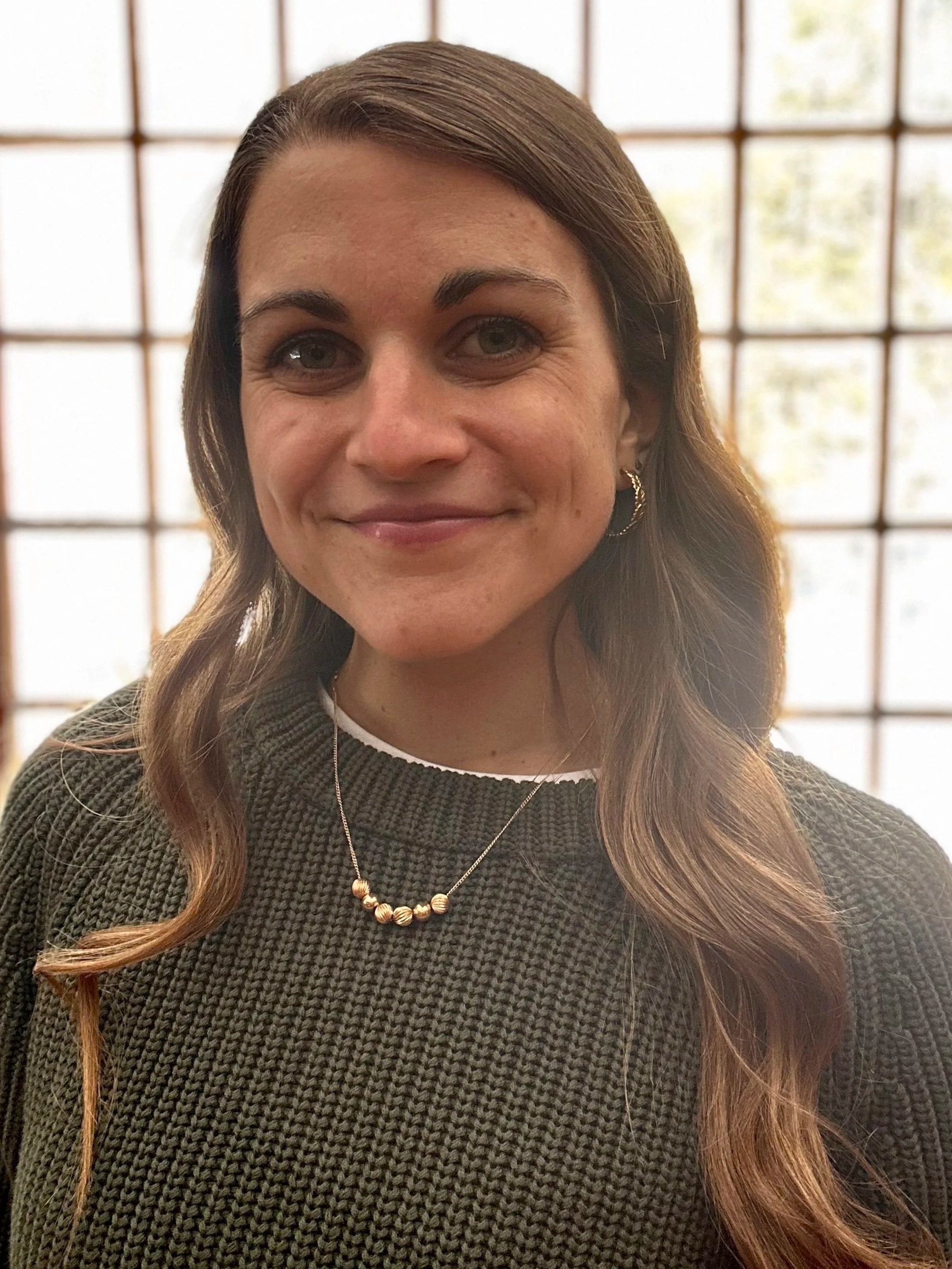 Close-up of a woman with long, wavy brown hair wearing a dark-colored sweater, gold earrings, and a gold bead necklace, standing in front of a window grid with a blurred outdoor background.