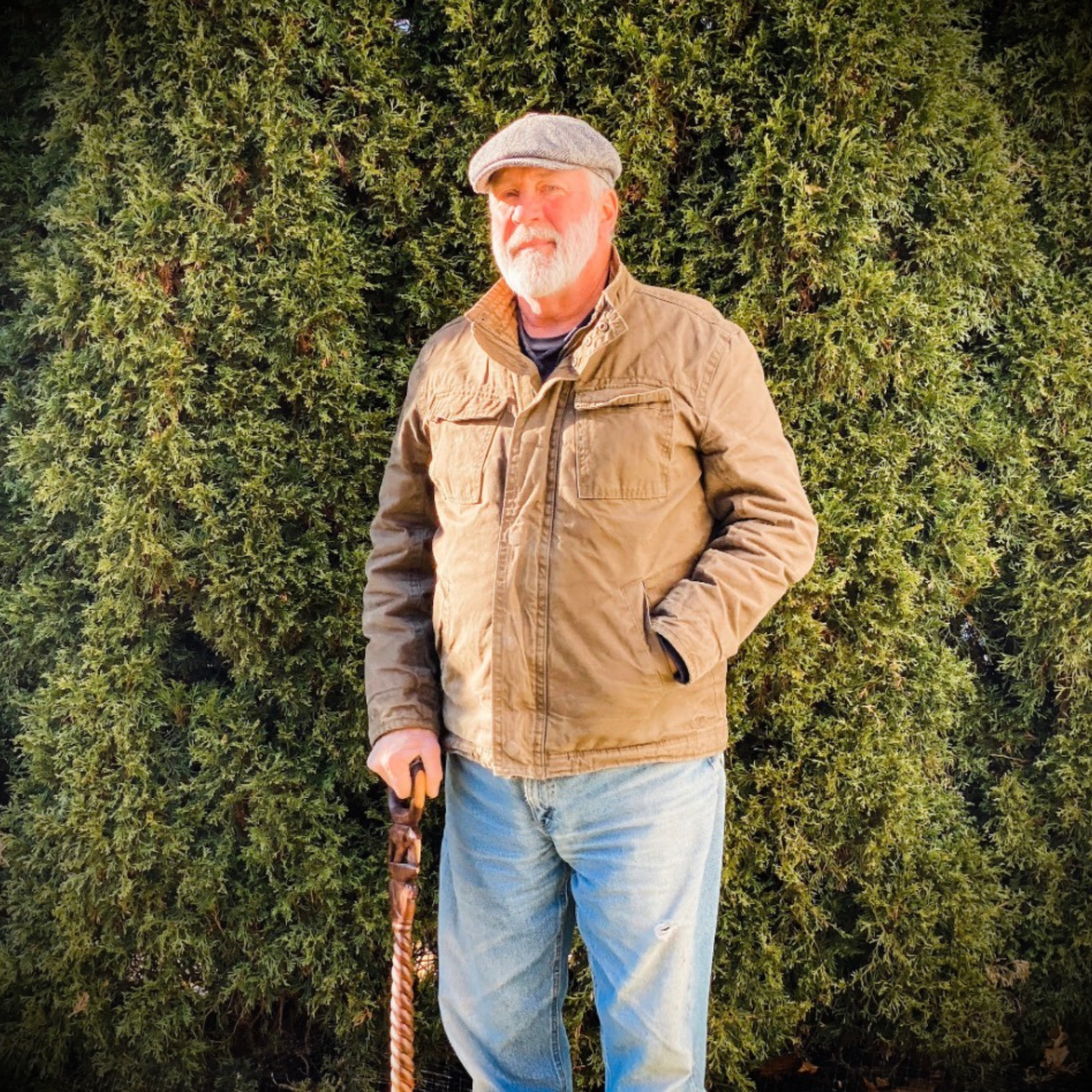 An elderly man with a white beard and gray hair wearing a gray flat cap, brown jacket, and jeans standing outdoors in front of green bushes.