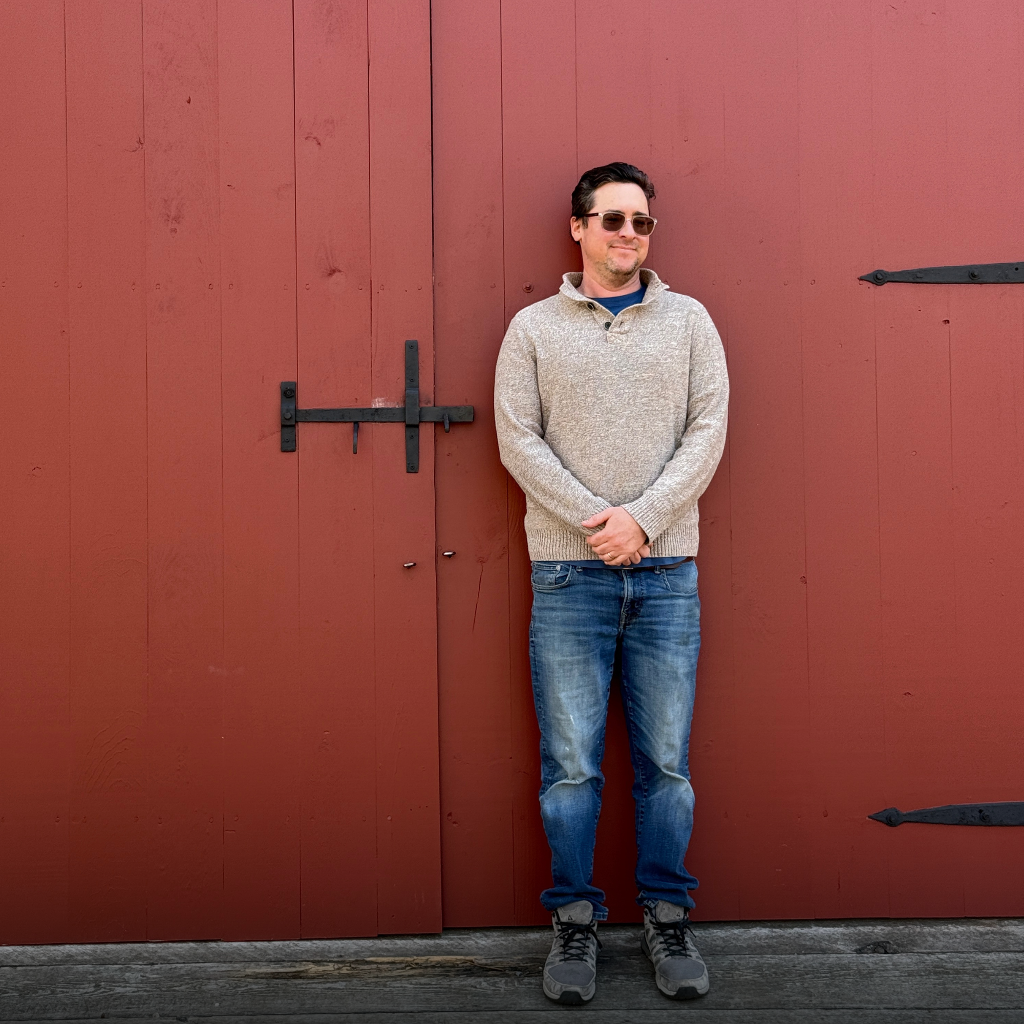 A man wearing sunglasses, a gray sweater, jeans, and hiking boots, standing with arms crossed in front of a large red barn door with black hardware, on a wooden surface.