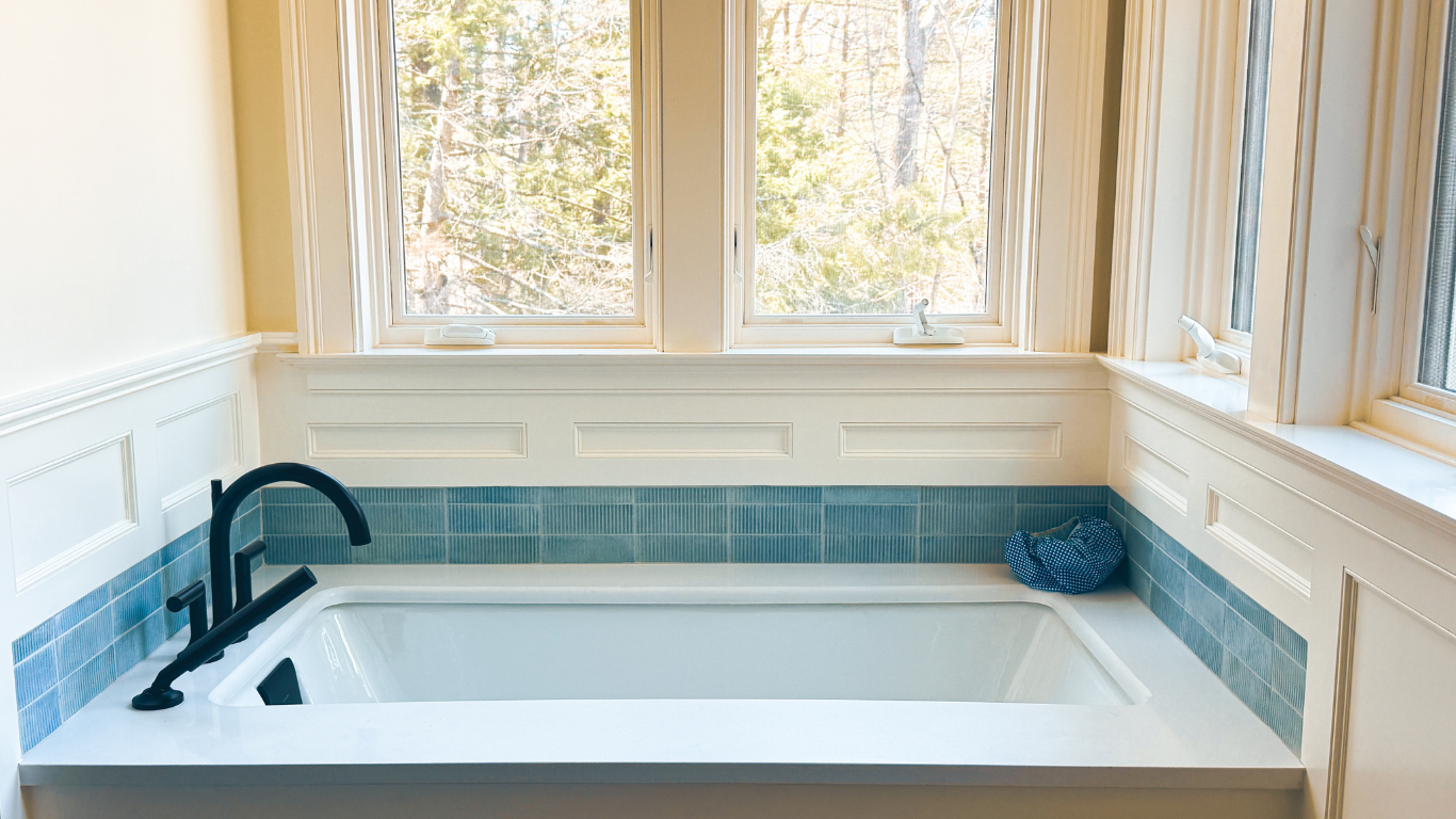 A bathroom with a large white bathtub beneath three windows. The windows show trees outside, and the bathtub has a black faucet and a small blue pillow on the corner.