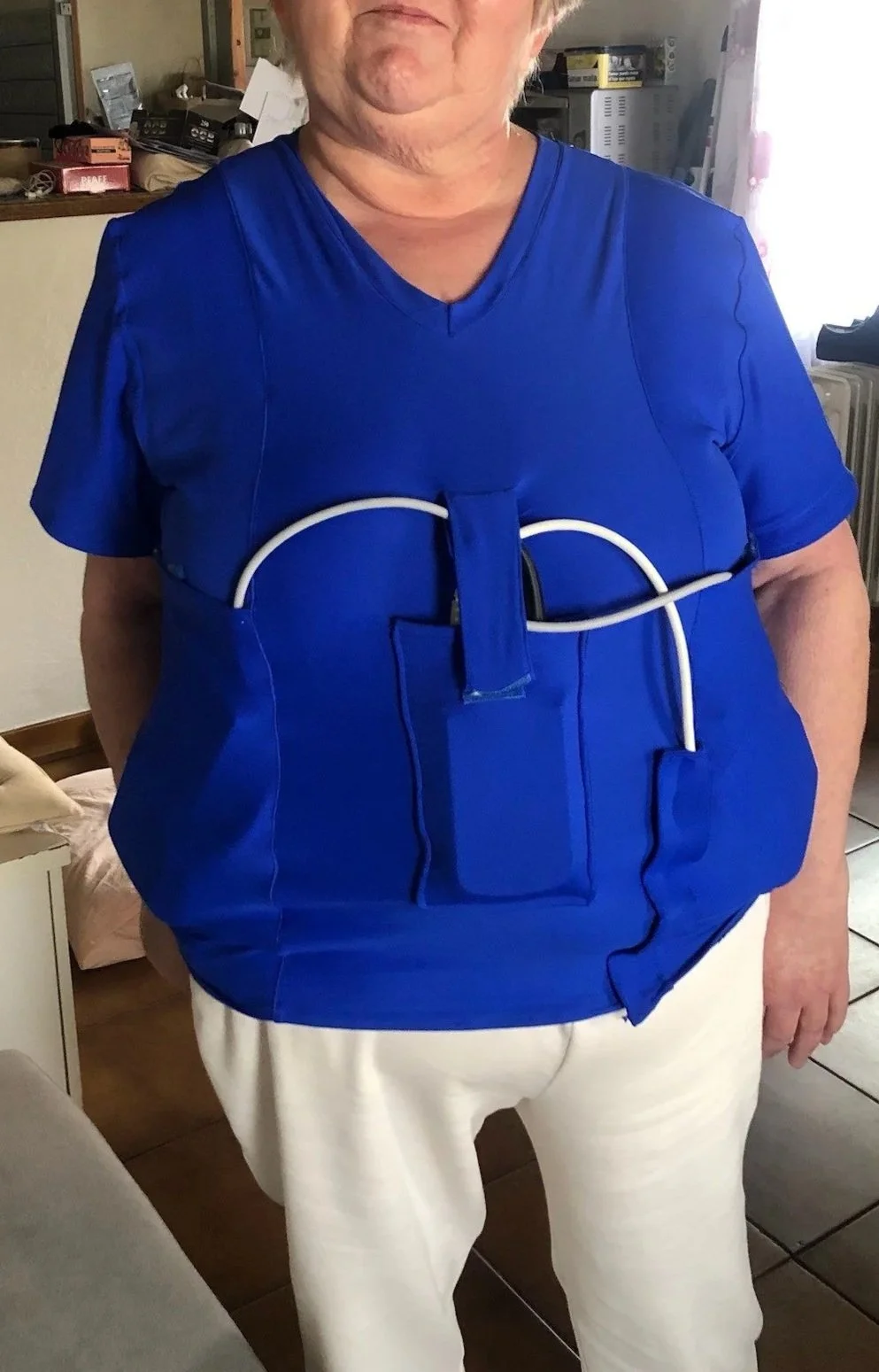 Une personne portant un vêtement bleu avec un dispositif médical attaché à l'avant, dans un intérieur domestique.