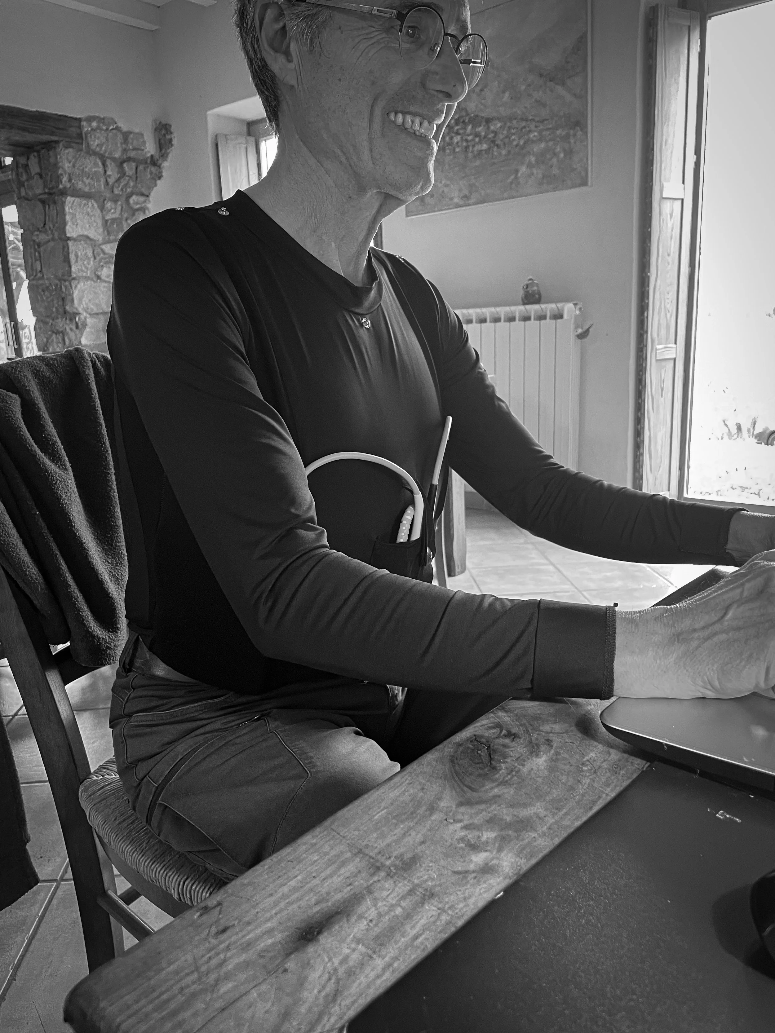 Un homme souriant avec des lunettes, assis à une table près d'une fenêtre, travaillant sur un ordinateur dans une pièce lumineuse.