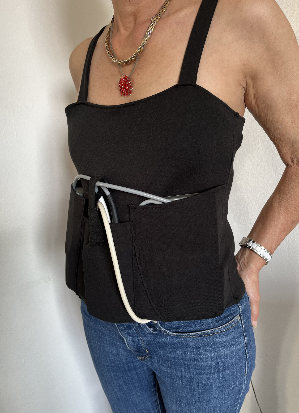 Une femme porte un haut noir et un portefeuille avec des câbles électroniques visibles, un collier orné d'une médaille rouge, une montre en argent, et des jeans bleus.