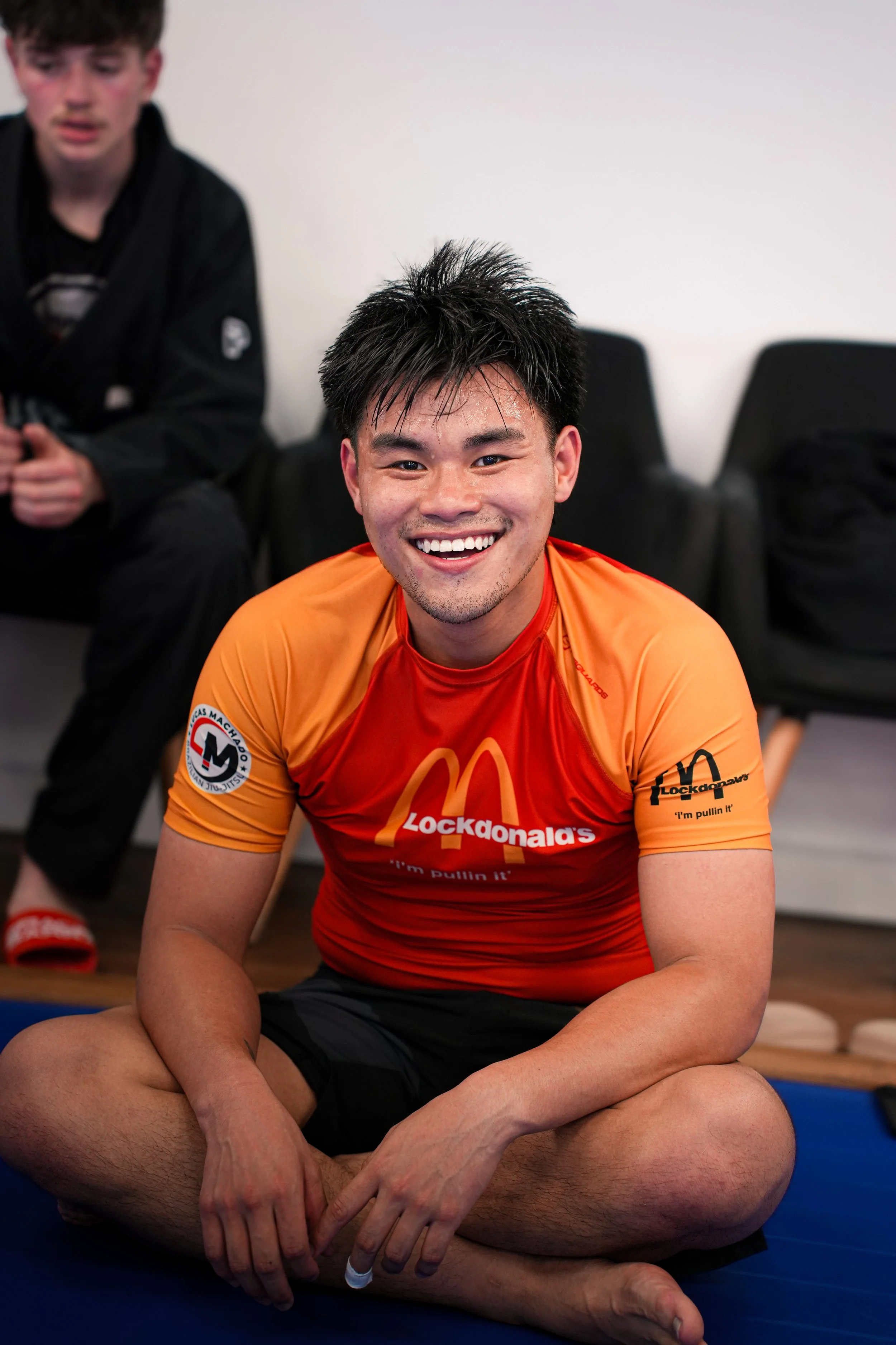 A smiling young man sitting cross-legged on a blue mat, wearing an orange sports shirt with McDonald's logos. In the background, another person in a black jiu-jitsu gi and a patch on his sleeve can be seen. The setting appears to be a martial arts or Brazilian jiu-jitsu gym.