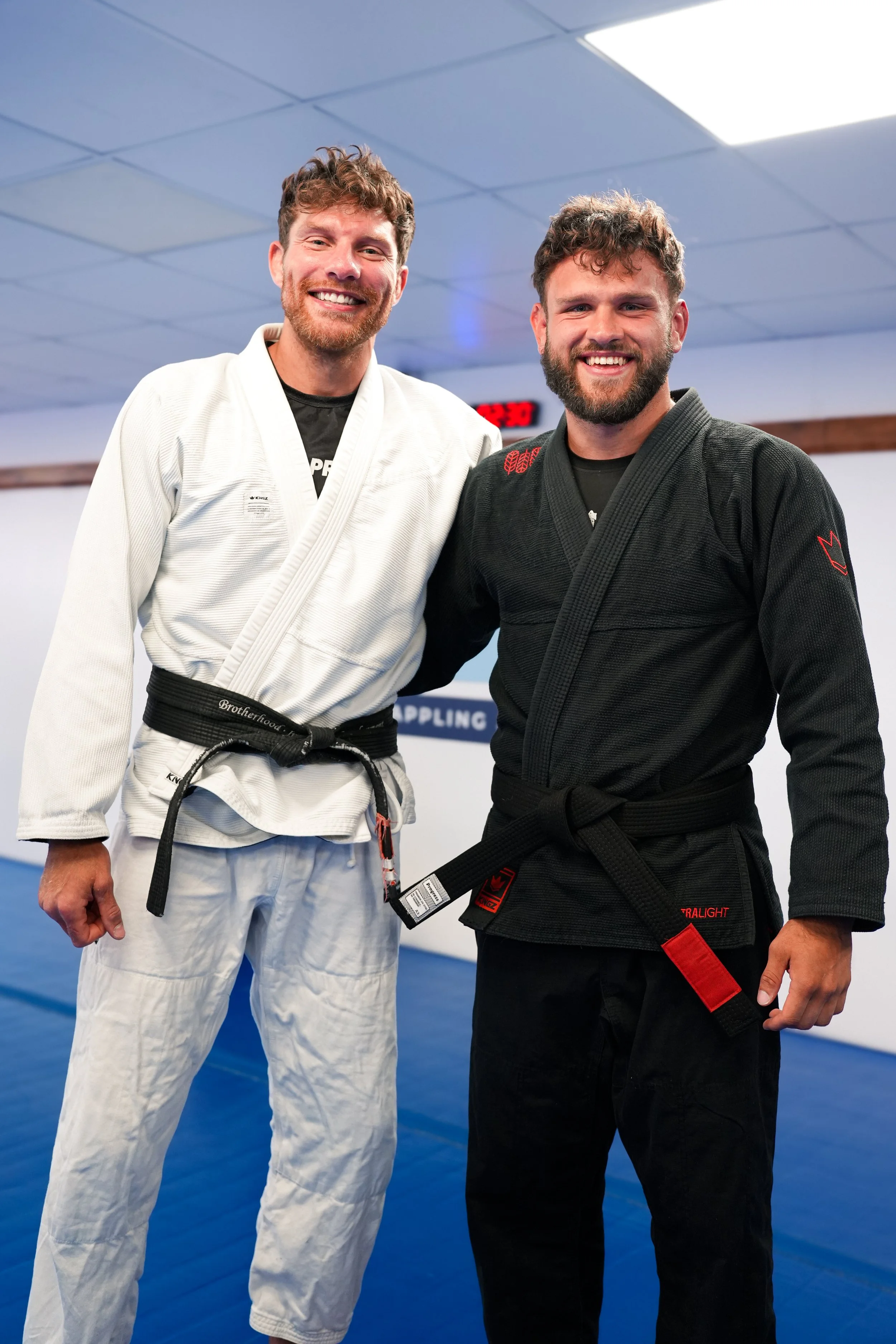 Two men in martial arts uniforms standing side by side and smiling indoors on a blue mat.
