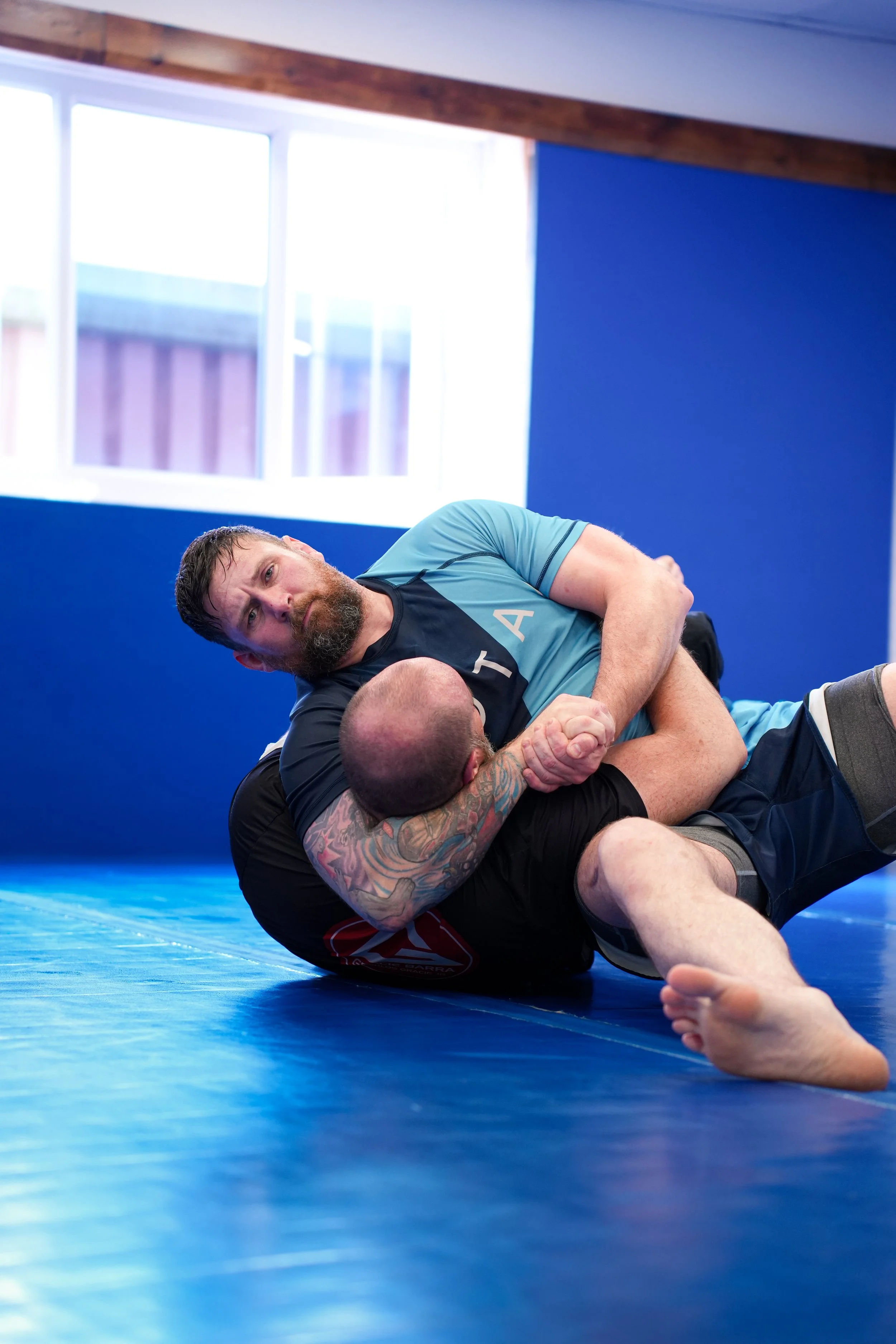 Two men practicing Brazilian Jiu-Jitsu on a blue mat inside a gym, engaged in a grappling move with one man on top and the other on the ground. Bright window light streams into the room.