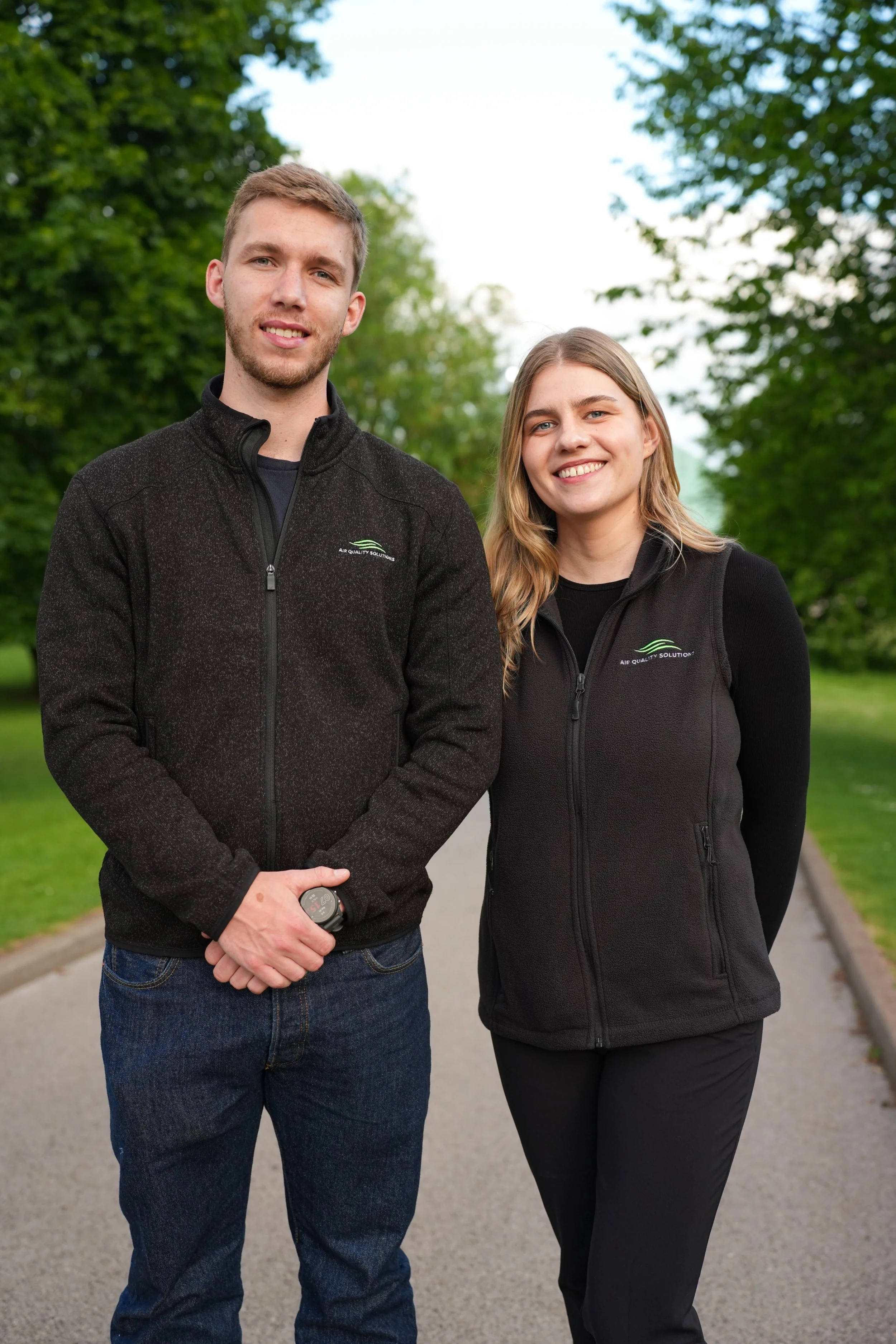 A young man and woman standing outdoors on a paved path surrounded by green trees, both smiling at the camera, wearing black jackets with a logo that says 'Air Quality Solutions'.