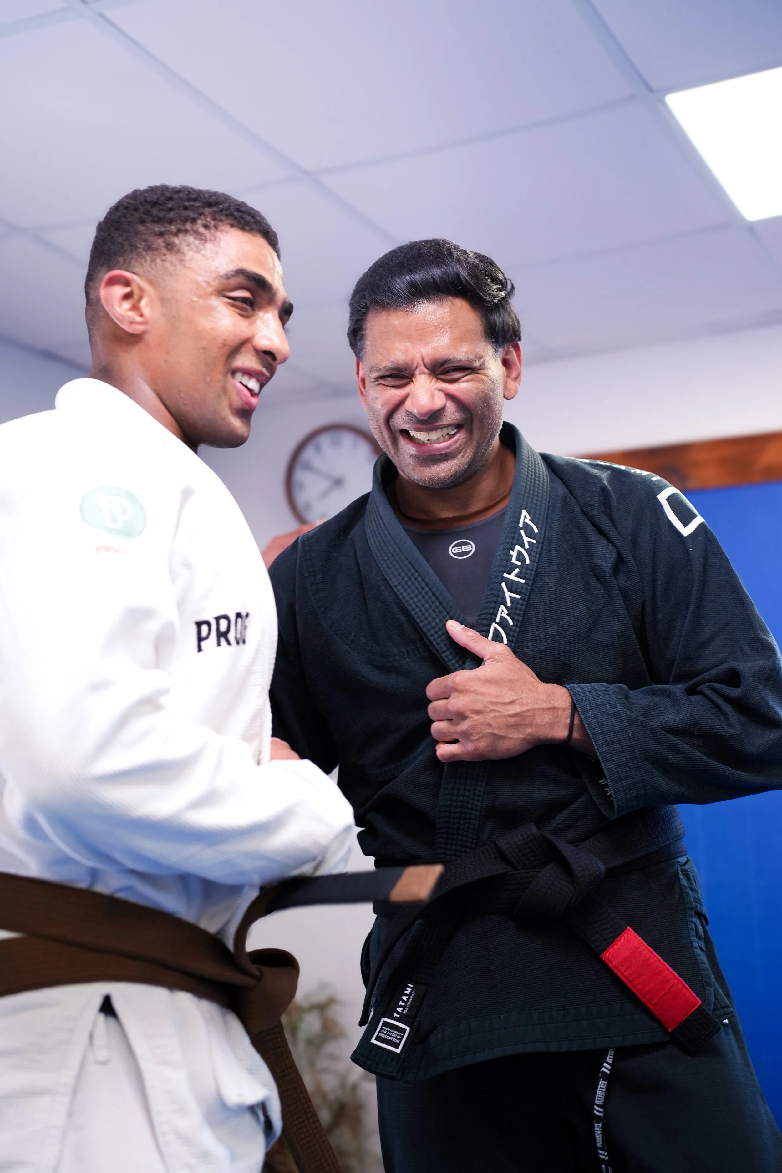 Two men in martial arts uniforms engaging in a martial arts practice or sparring session, smiling and showing camaraderie in an indoor training facility.