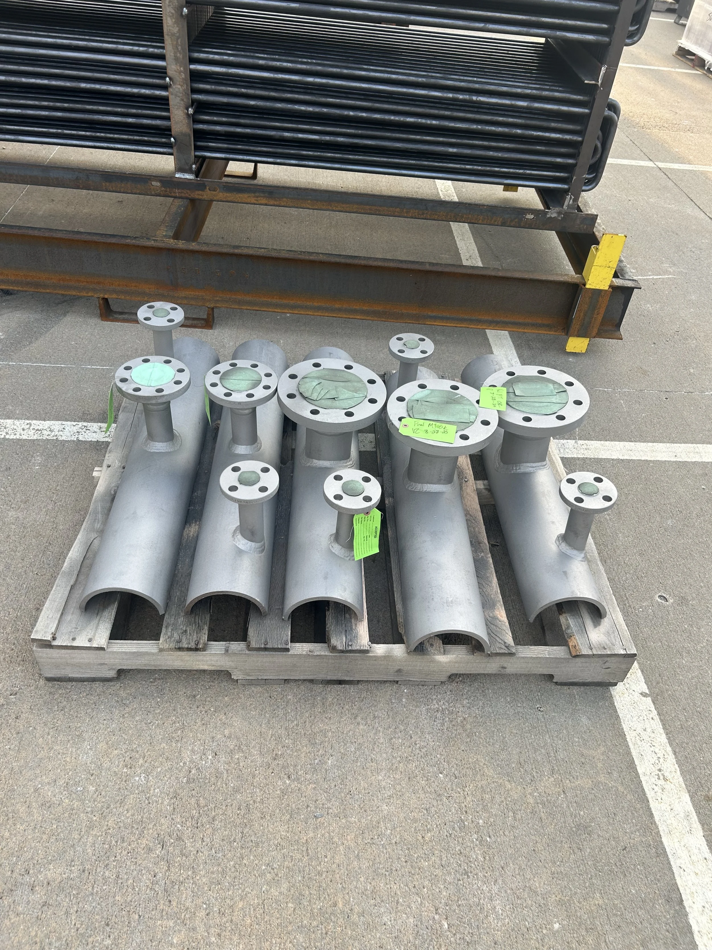 Various metal pipes and flanges on a wooden pallet, stored on an outdoor pavement.