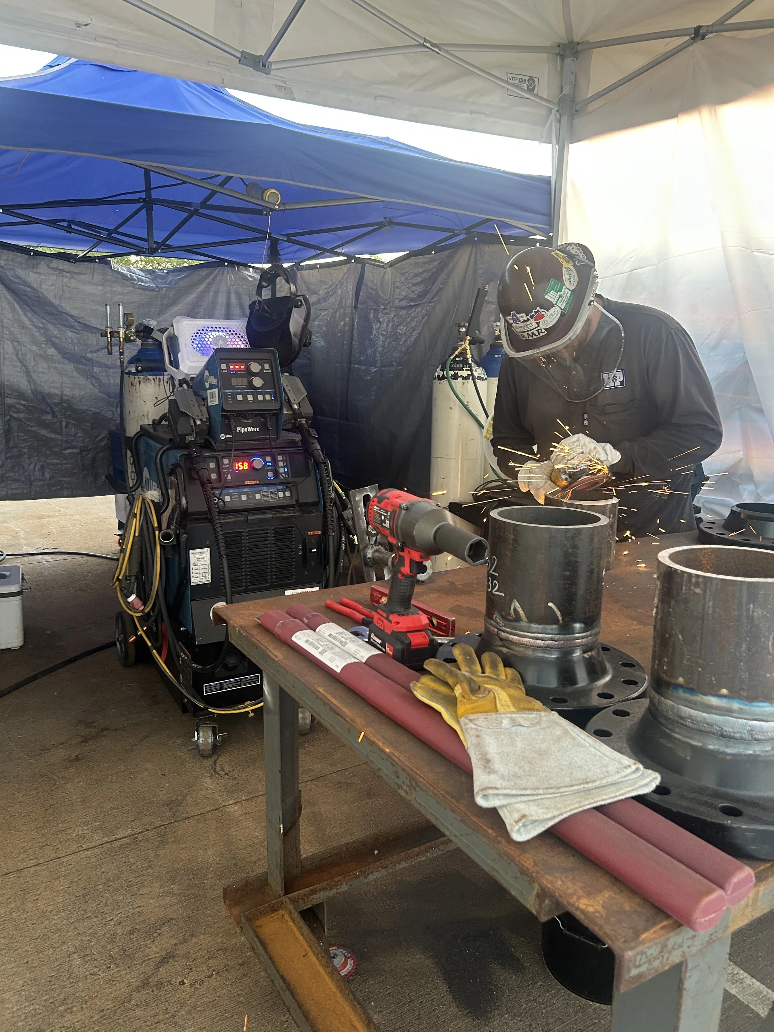 A person welding metal pipes at a work station inside a tent, with welding equipment, gloves, and tools on a table.