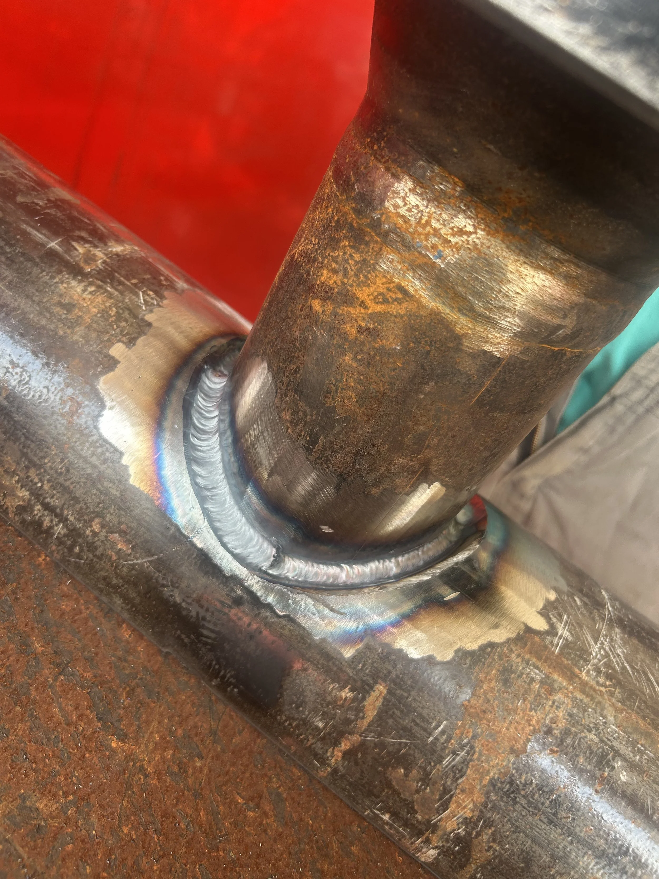 Close-up of a welded metal joint showing a cylindrical metal tube attached to a flat metal plate, with visible weld beads and heat discoloration around the weld area.