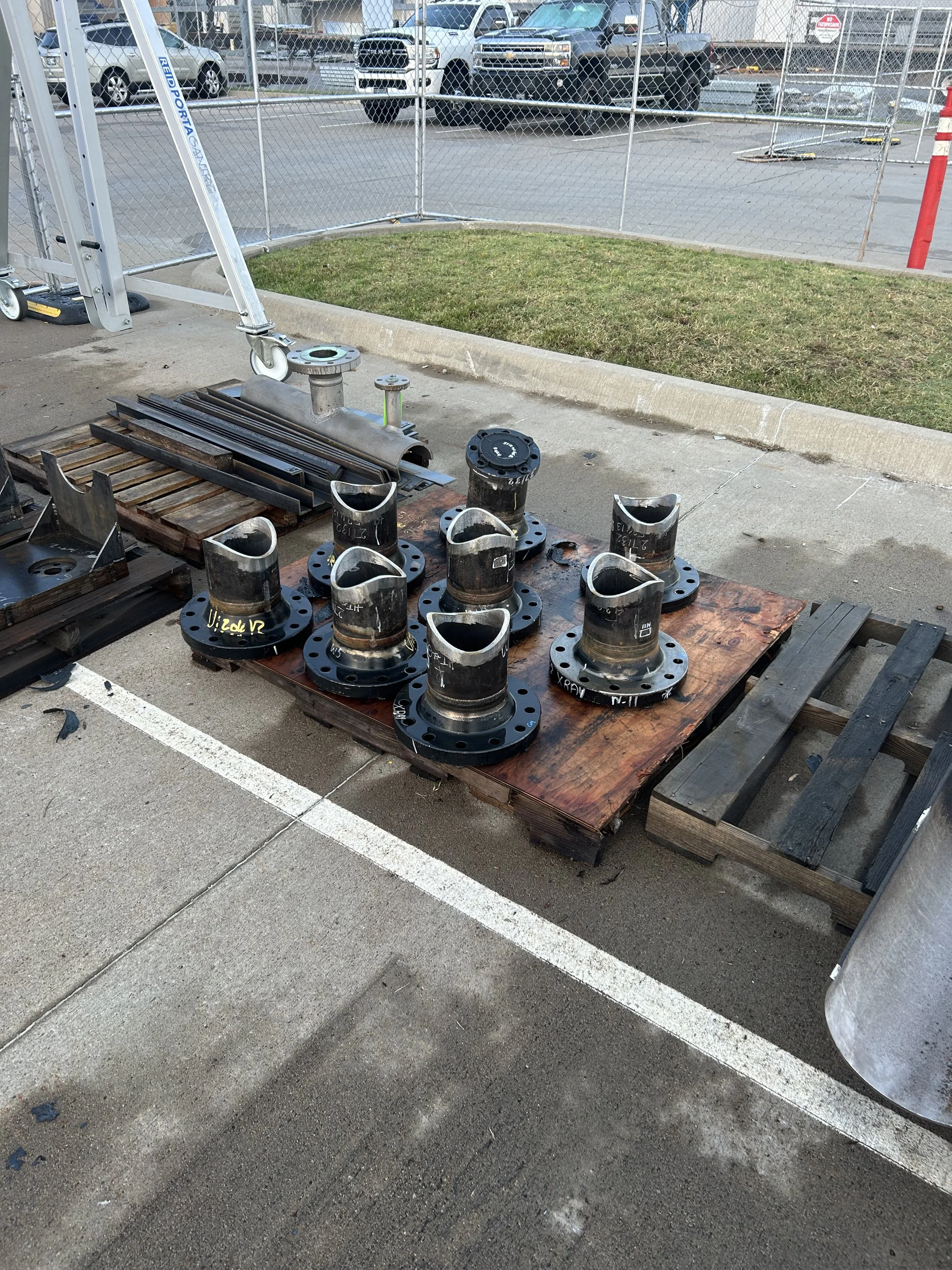 Metal pipe fittings with flanges, pipes and metal parts arranged on wooden pallets outside, near a construction site with a fence and parked cars in the background.