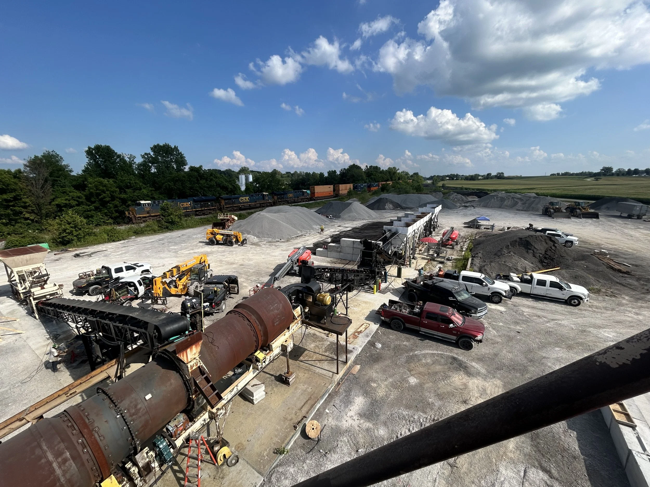 Construction site with various machinery, piles of gravel and dirt, parked trucks and equipment, and a train in the background under a partly cloudy sky.