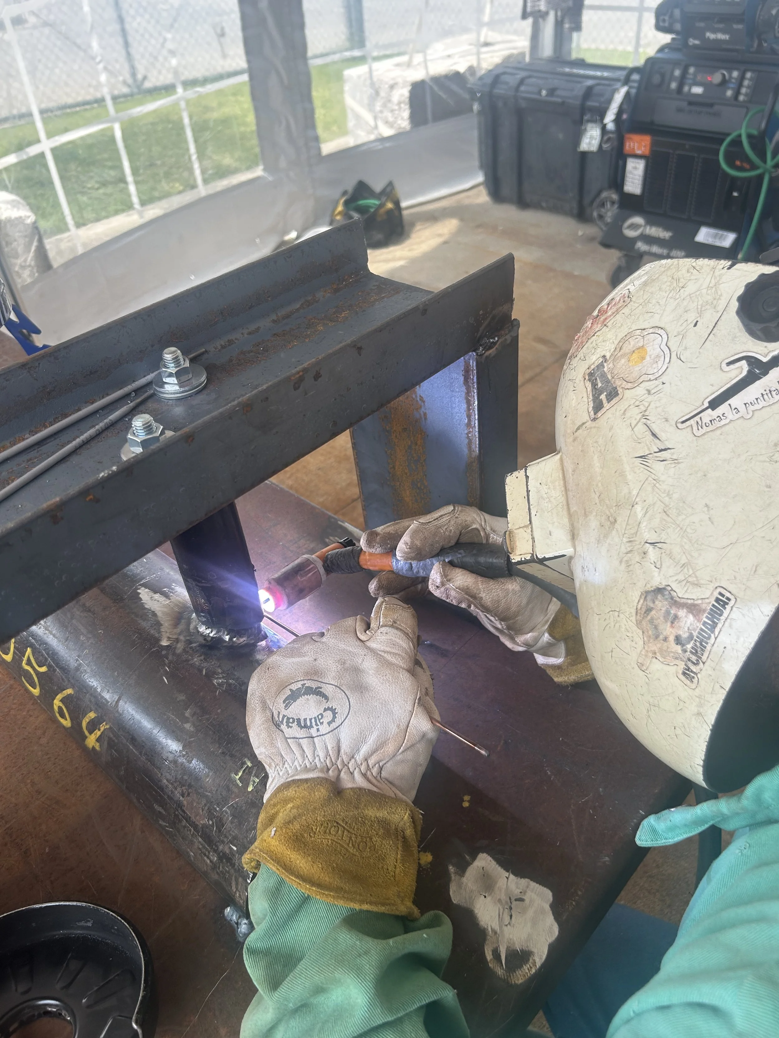 A person welding a metal pipe with a welding mask and gloves at a workbench outdoors.