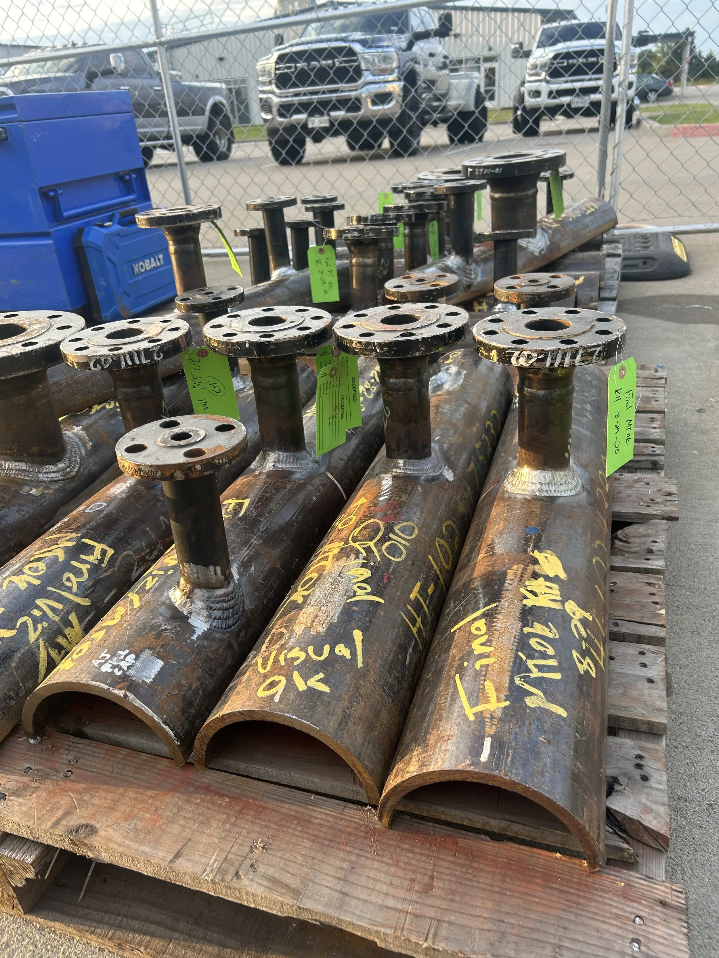 Large rusted metal pipes with flanges, labeled with yellow and white markings, placed on a wooden pallet outdoors. In the background, there are parked trucks and a chain-link fence.