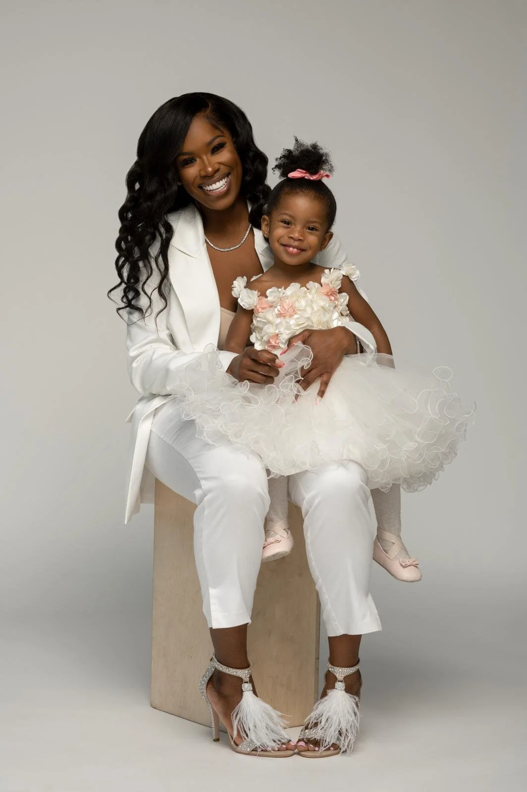 A woman and a young girl are sitting together, both smiling. The woman has long, wavy black hair and is dressed in a white suit with high-heeled, feathered shoes. The girl is wearing a white dress with pink flowers on the top, a matching pink bow in her hair, and pink shoes with bows. The background is plain and light-colored.