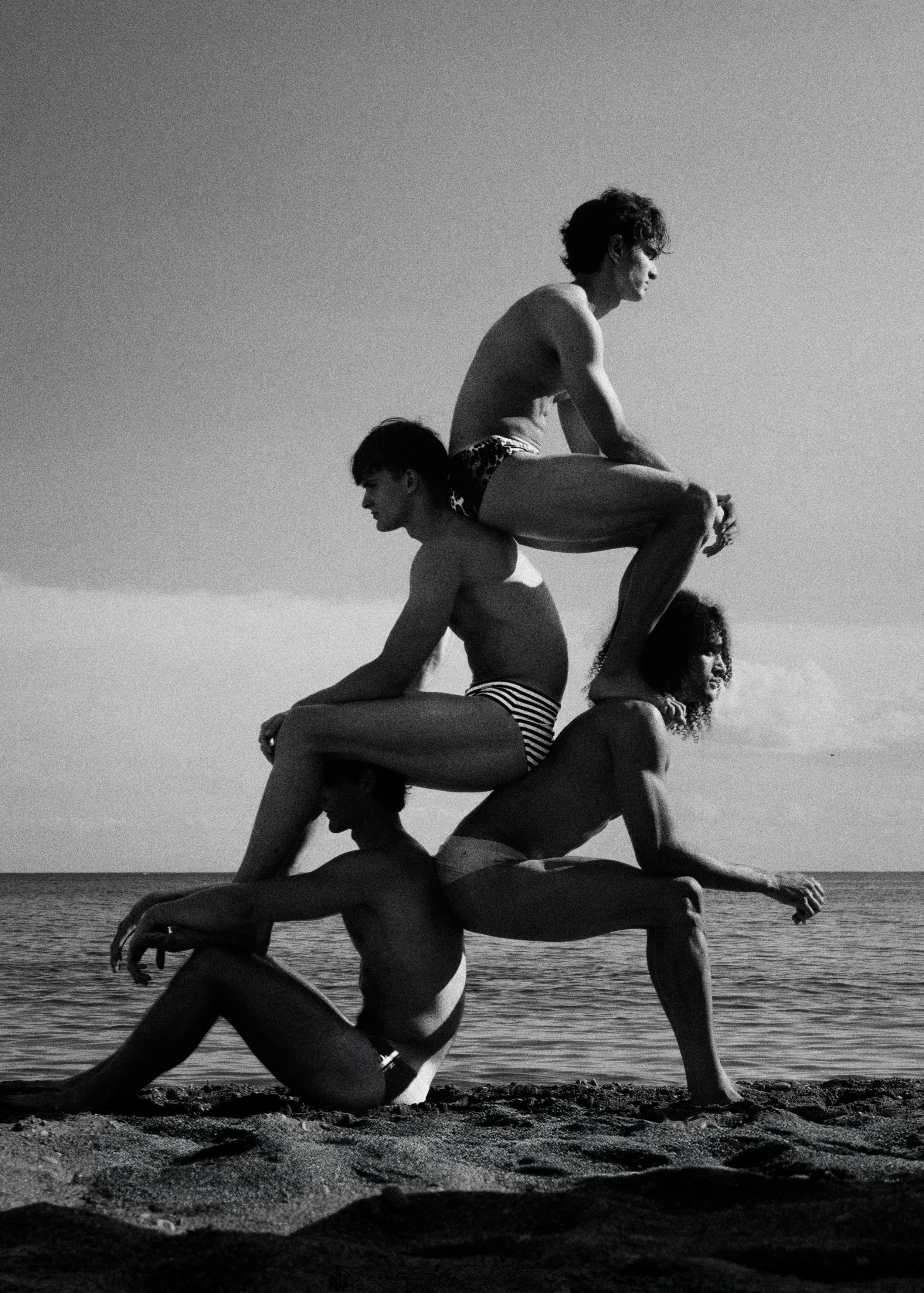 Four people stacked on each other's backs on a beach, creating a human tower, with the ocean and sky in the background.  By Nick Merzetti