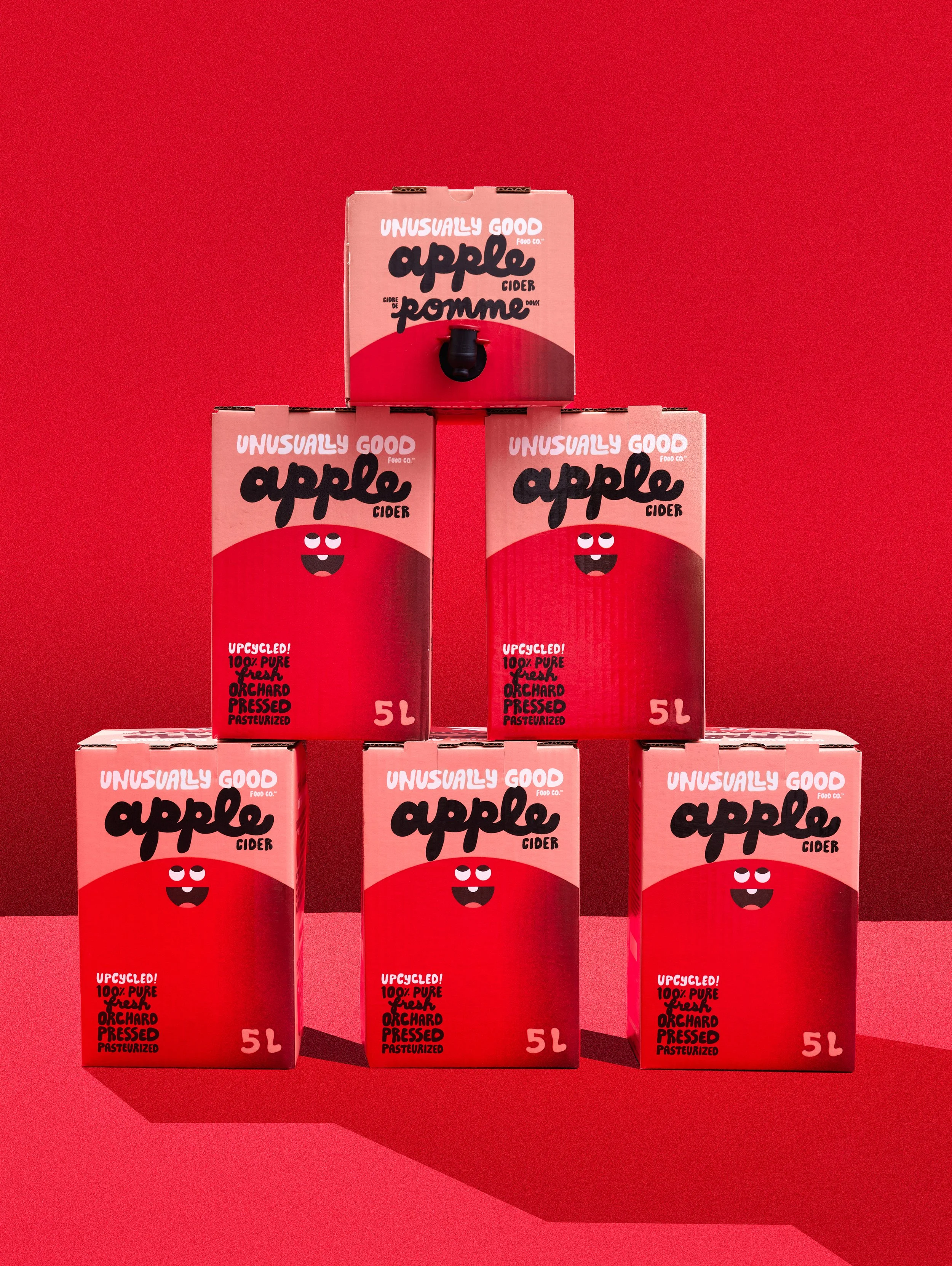 Five red boxes of apple cider arranged in a pyramid shape on a red background. The boxes feature a cartoon apple face and text indicating they are upcycled, 100% pure, orchard pressed, pasteurized cider, each holding 5 liters.  By Nick Merzetti