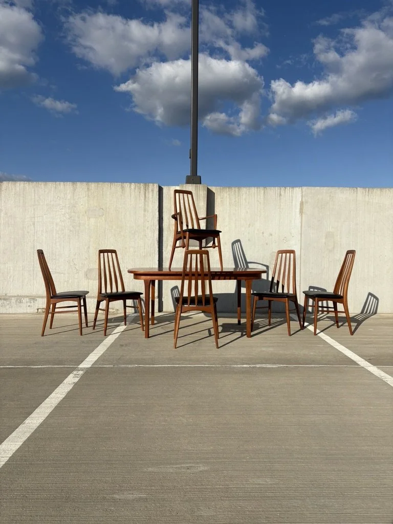 A group of Niels Koefeld "Eva" chairs arranged around a danish dining table on a parking deck, with a concrete wall and a blue sky with clouds in the background.