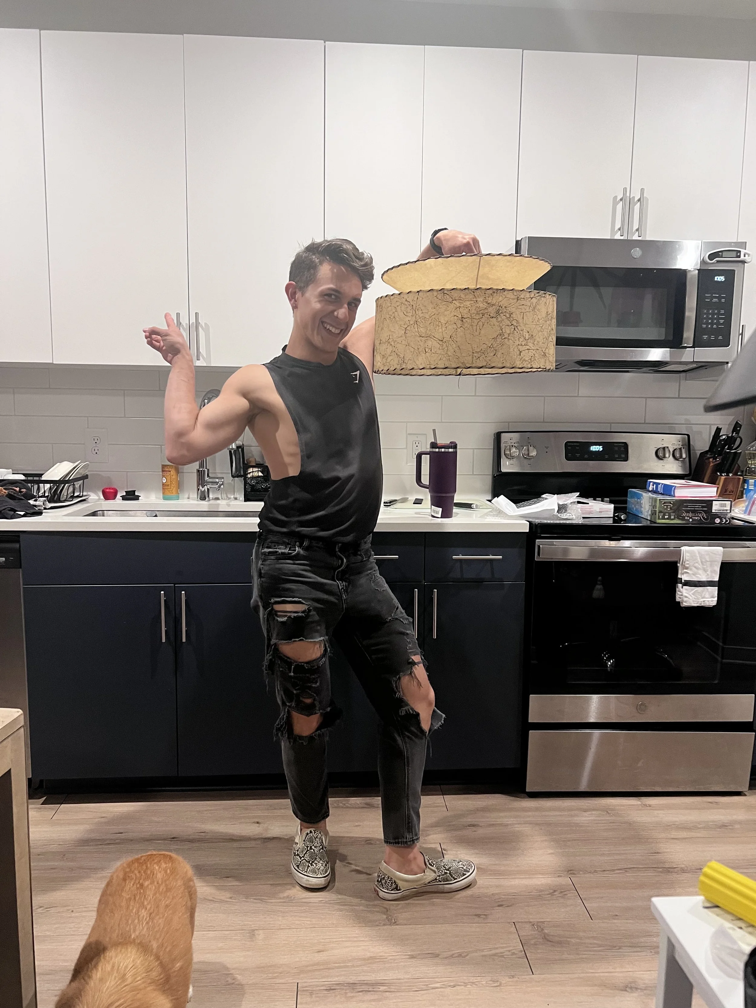 A young man with short dark hair, wearing a sleeveless black shirt, ripped black jeans, and snake-print shoes, is in a kitchen holding a large, atomic two-tier lamp shade, above his head with one hand, smiling at the camera.