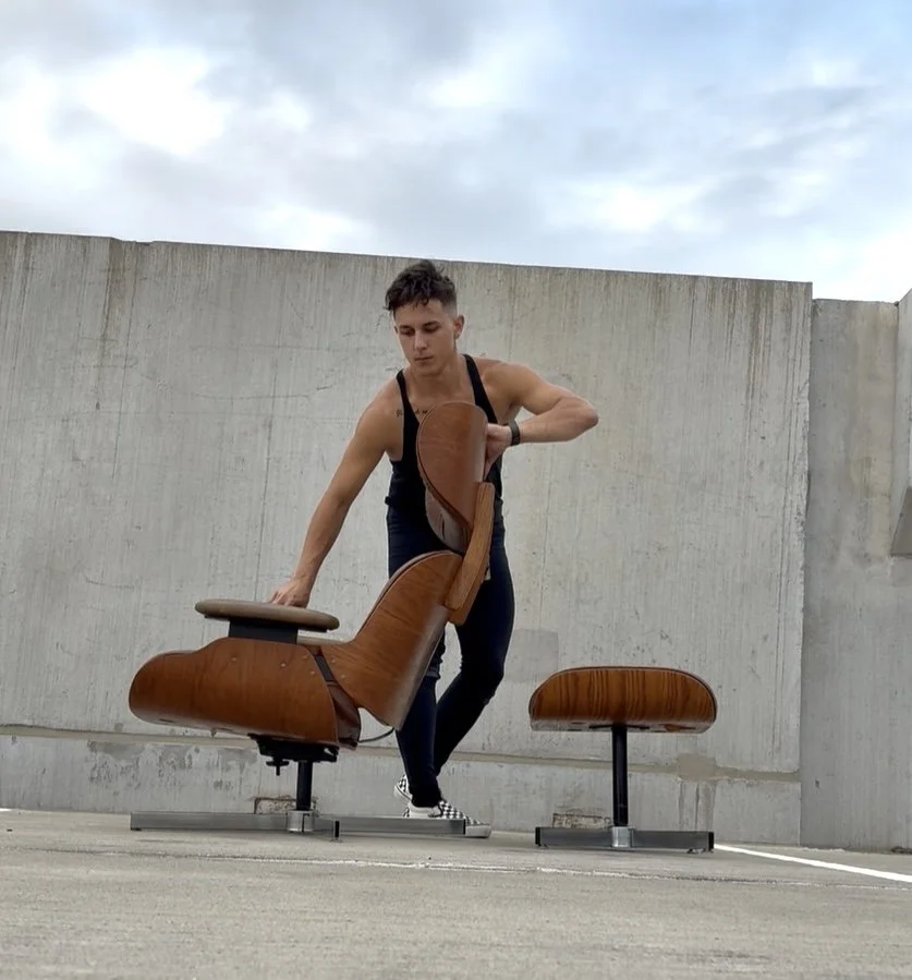 A young man adjusting an Eames style lounge chair on a rooftop parking lot with a concrete wall and cloudy sky in the background.
