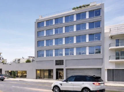 A daytime photograph of a four-story apartment building, with a white car driving past it.