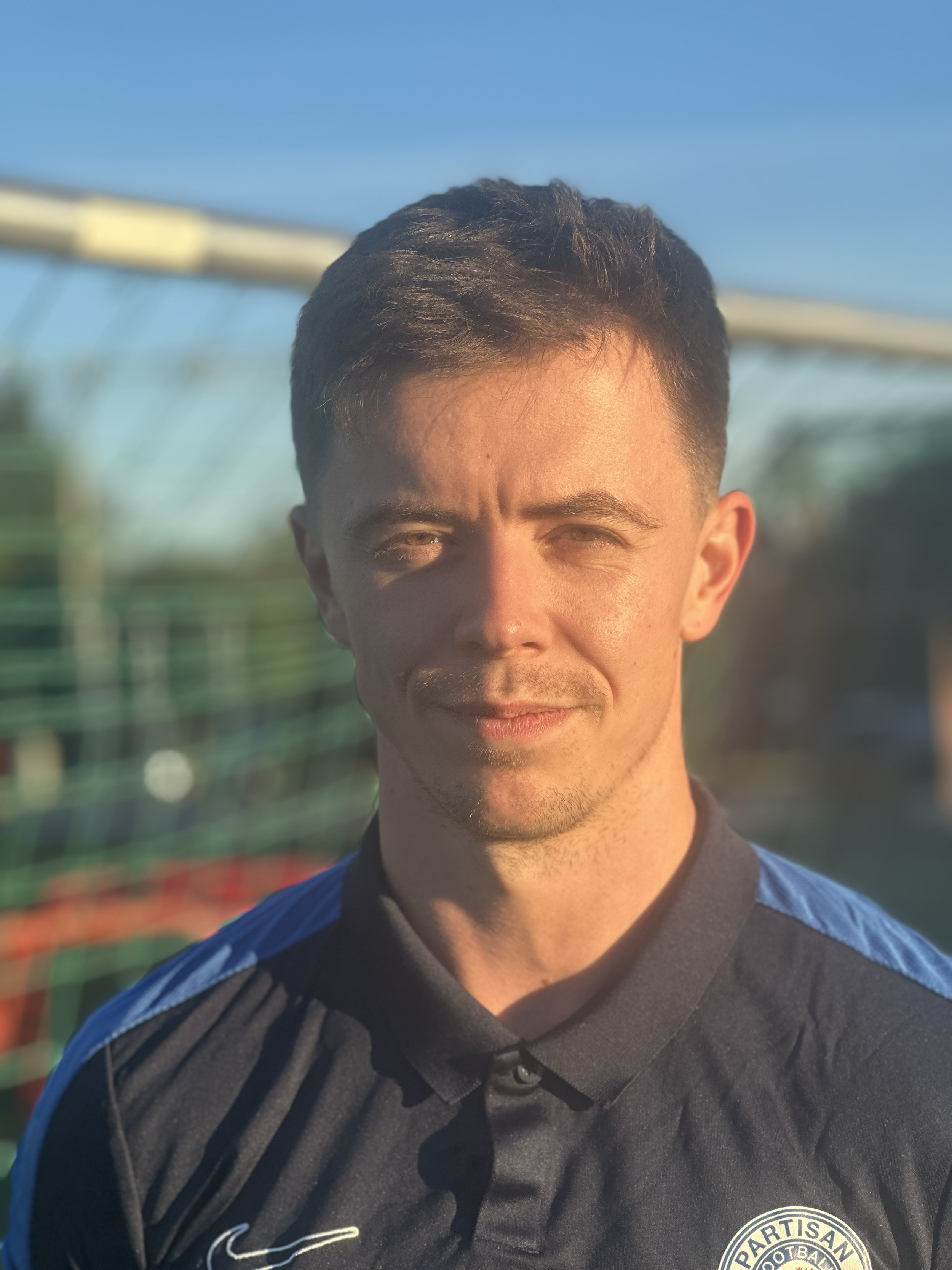 Young man with short dark hair and light skin, wearing a navy jersey with a Partisan football club logo, standing outdoors near a fence under a blue sky.
