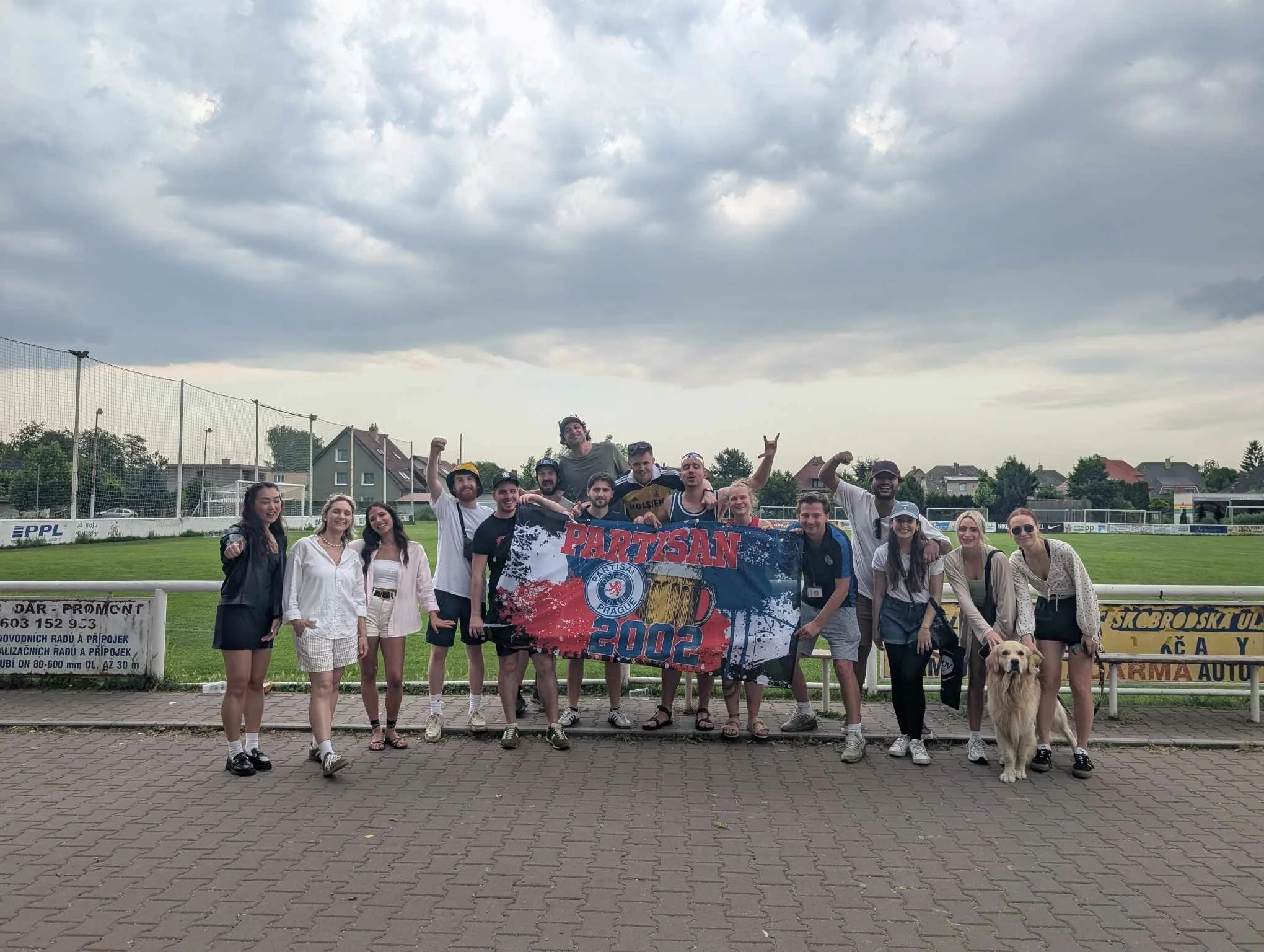 Group of people standing together on a sports field, holding a banner that reads "Partisan 2002." They are smiling and celebrating, with some making celebratory gestures. There is a dog in front of the group, and houses and a cloudy sky are visible i
