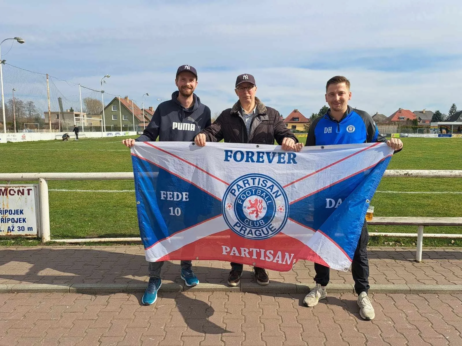 Three men holding a flag in front of a soccer field, with houses and a clear sky in the background.