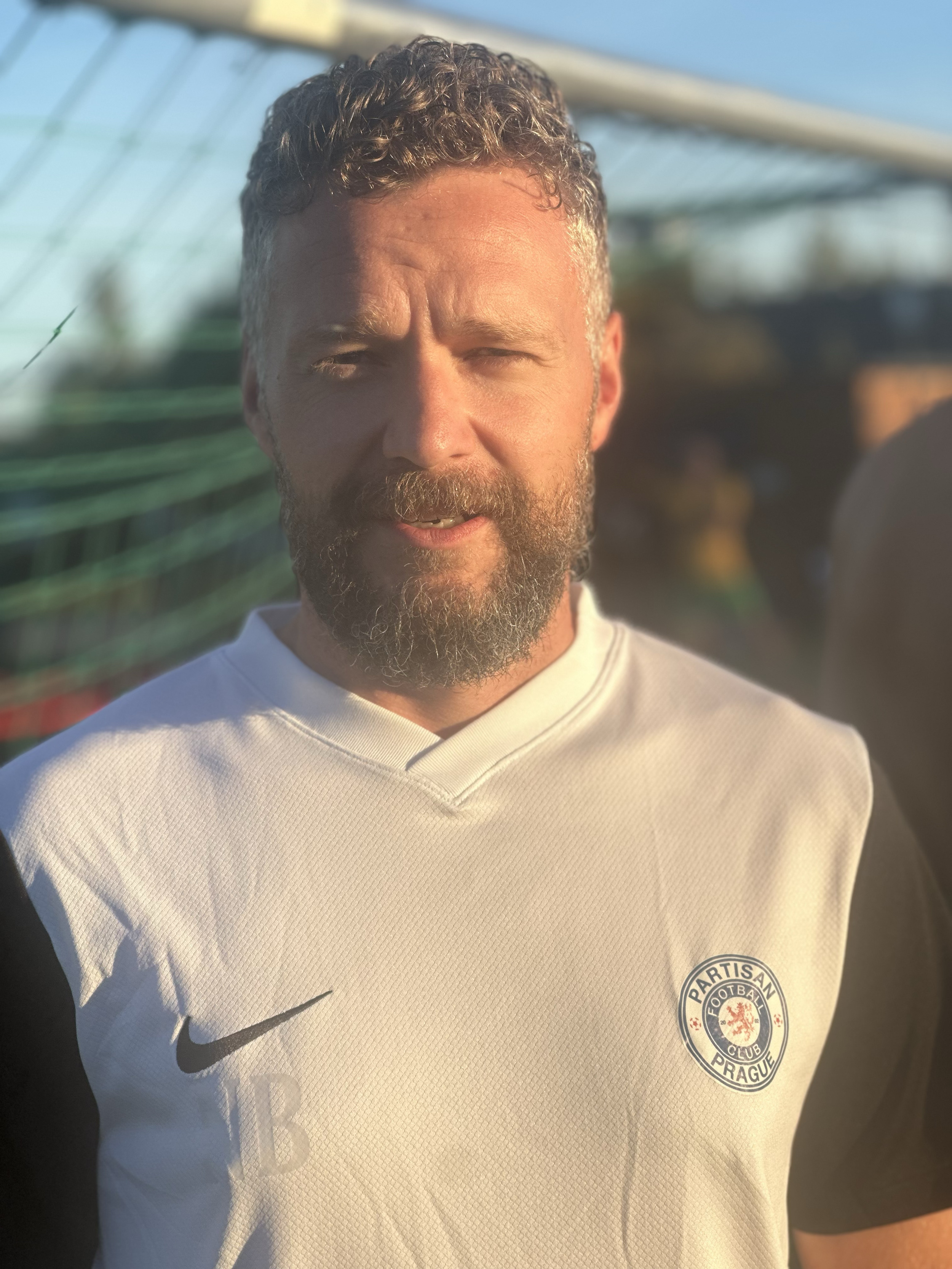 A man with curly hair and a beard wearing a white sports jersey with the logo of Partisan Football Club Prague. background features a sunny outdoor setting with a blurred fence or netting.