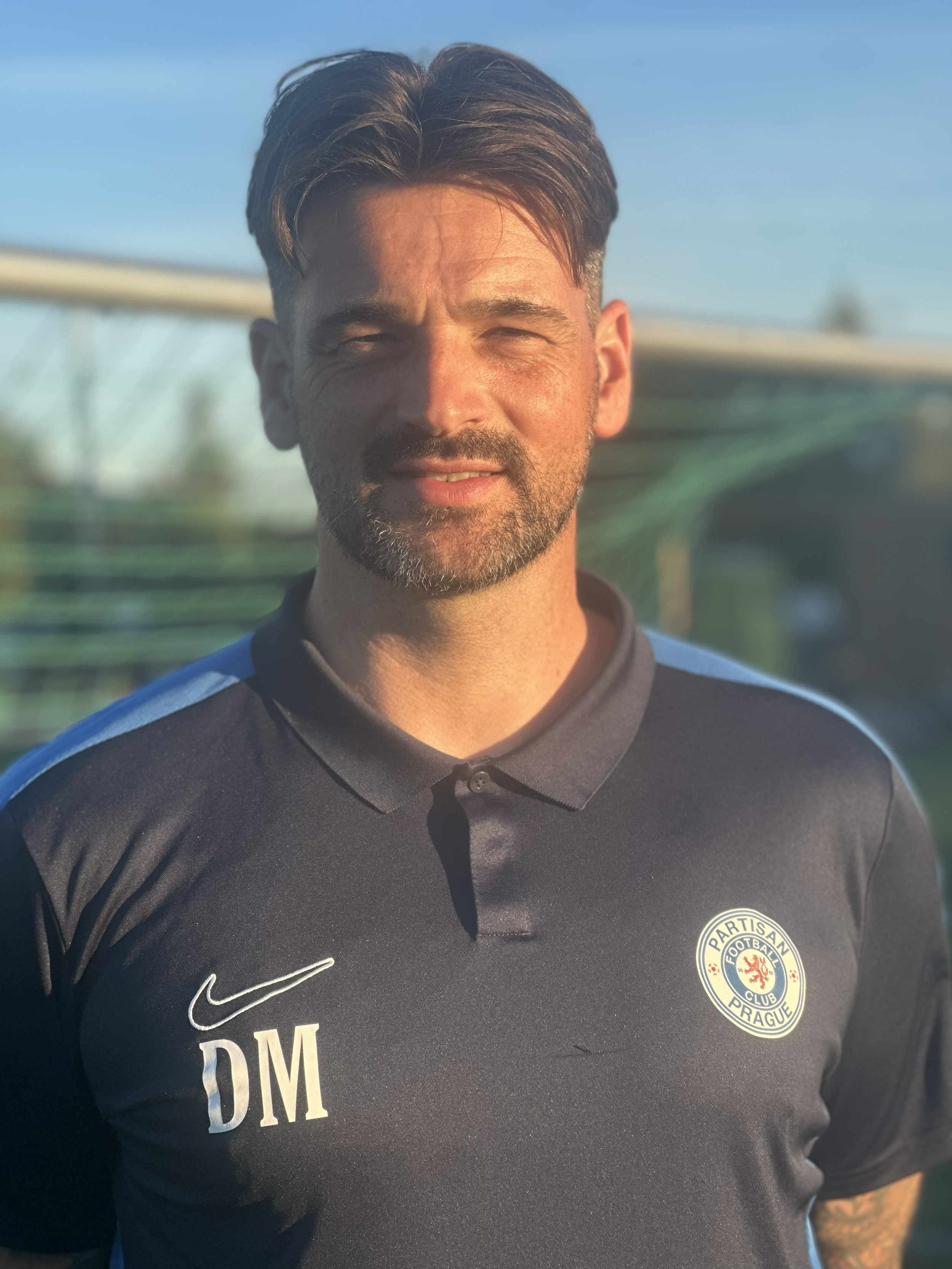 A man with dark hair and a beard wearing a black sports jersey with the logo of Partisan Prague football club and the initials DM, standing outdoors with a blurred sports field or stadium in the background.
