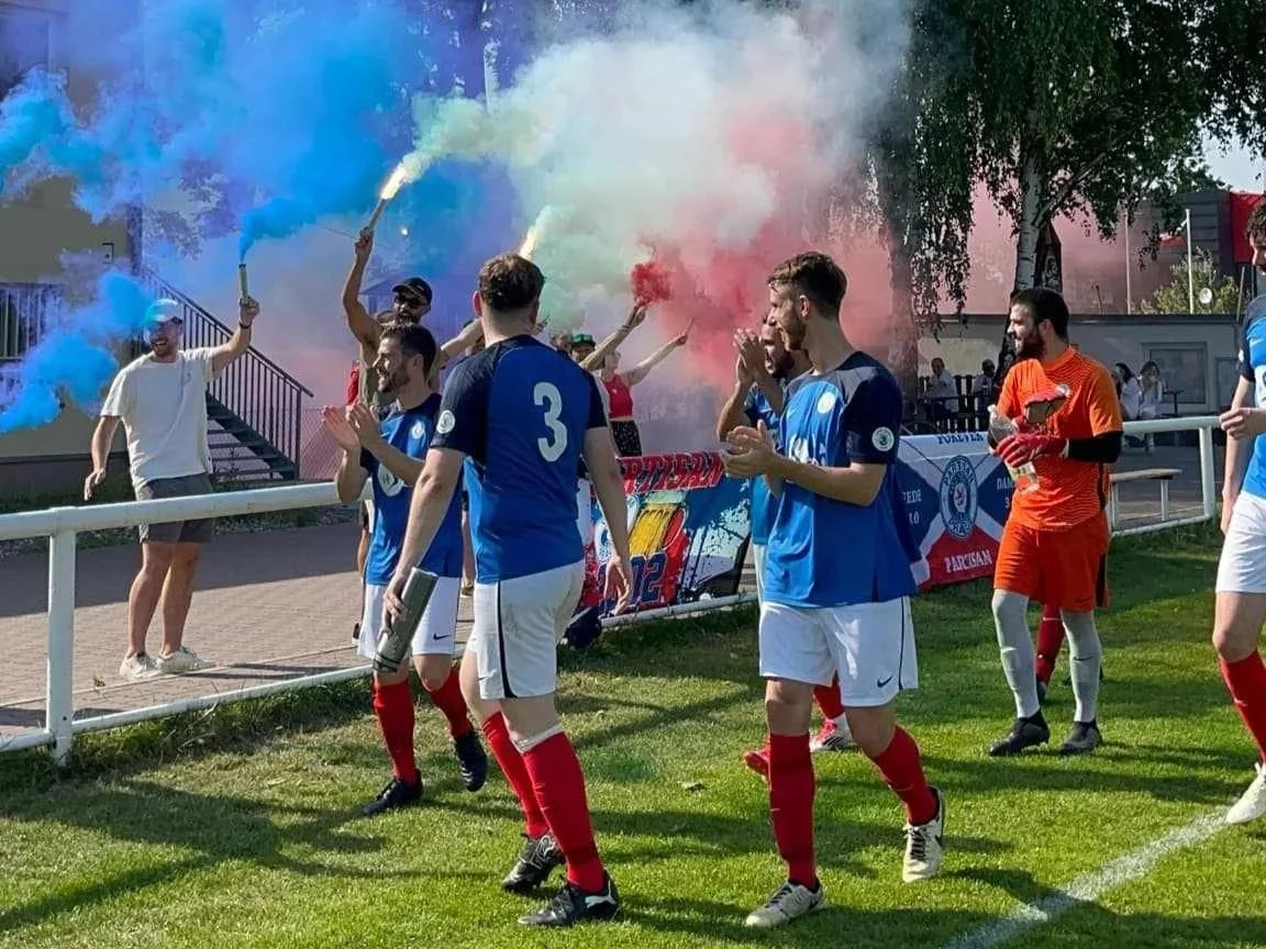 Soccer players celebrating on the field with smoke in the background, some fans are holding colorful smoke flares.