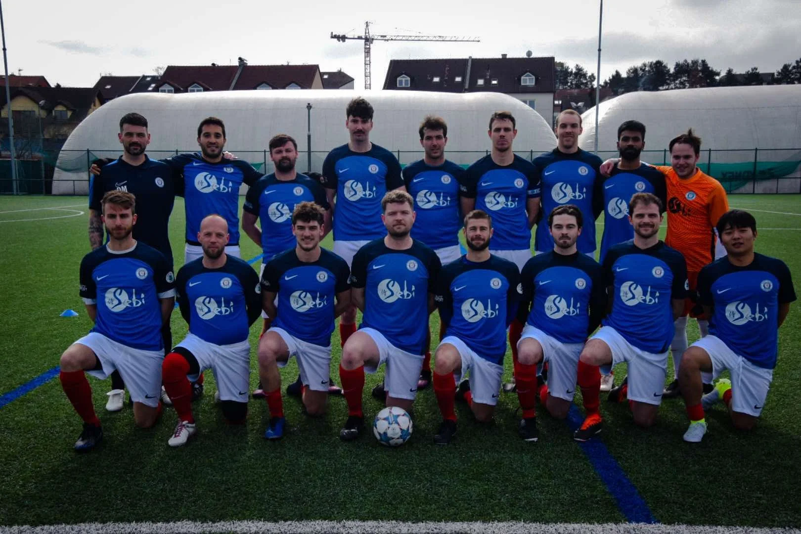 A group of soccer players in blue jerseys and white shorts posing on a soccer field, with team members kneeling in front and others standing behind, some with arms around each other.